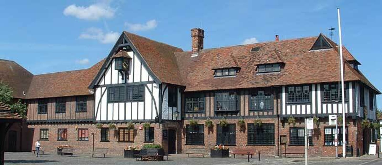 A historic building with a combination of timber and brick architecture, featuring a sloped roof and several windows. It's situated in a spacious courtyard under a clear blue sky. A few people and planters are visible in the foreground.