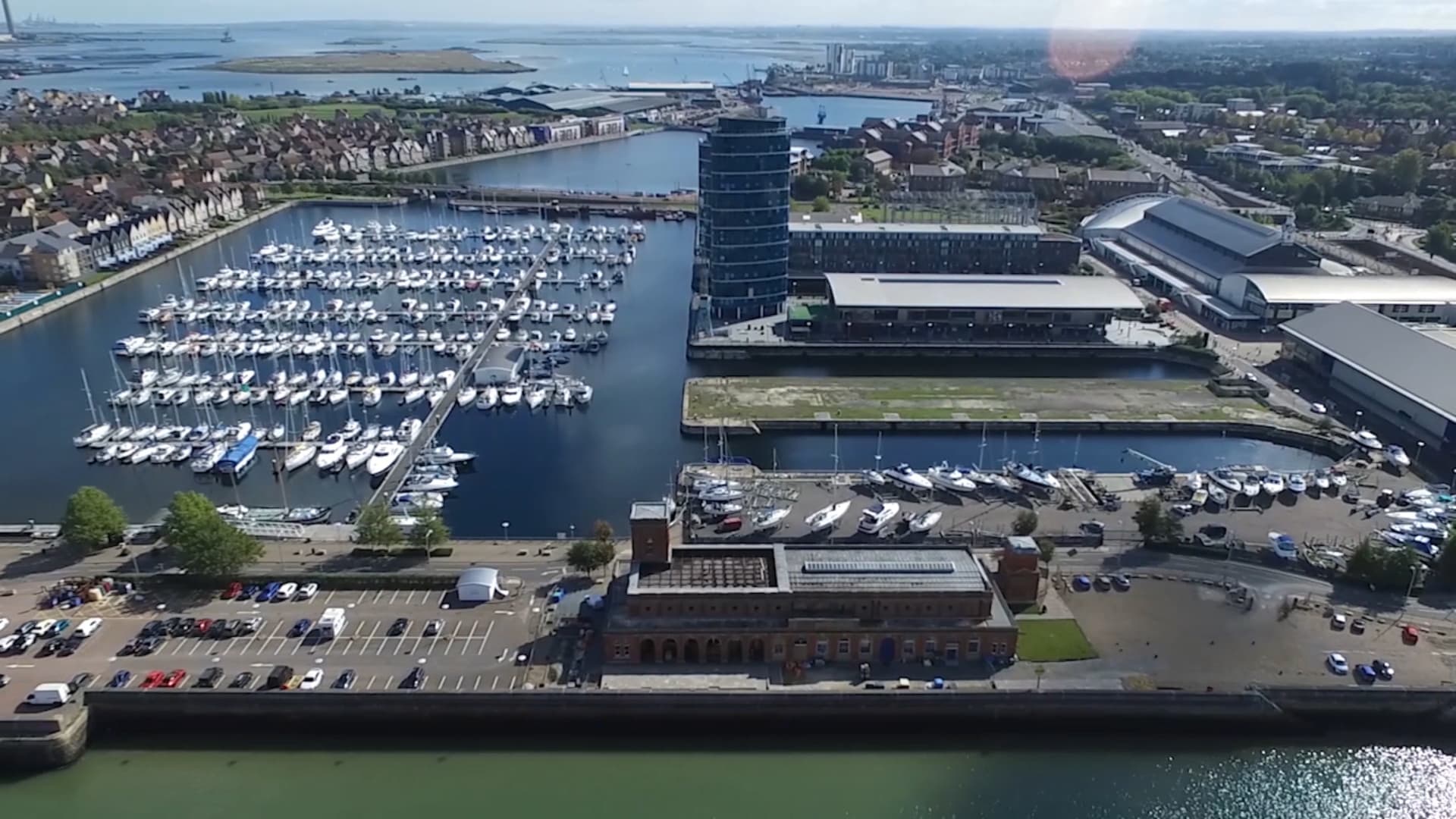 Aerial view of a coastal city featuring a marina filled with numerous boats. Buildings, a large circular structure, and a bridge are visible. The water is calm, with a distant backdrop of more water and land. Sunny weather highlights the scene.
