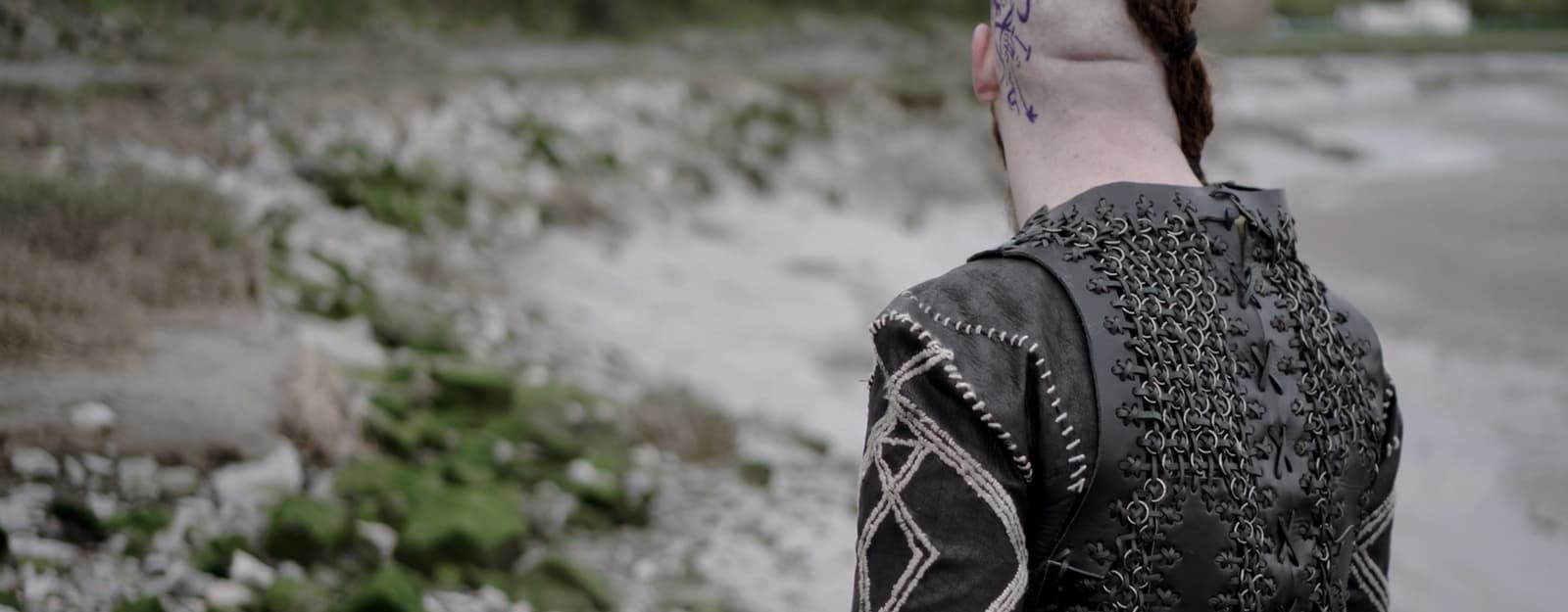 A person with braided hair and tattooed head is wearing a dark, intricately patterned jacket. They are standing on a rocky beach with patches of green moss, facing away from the camera.