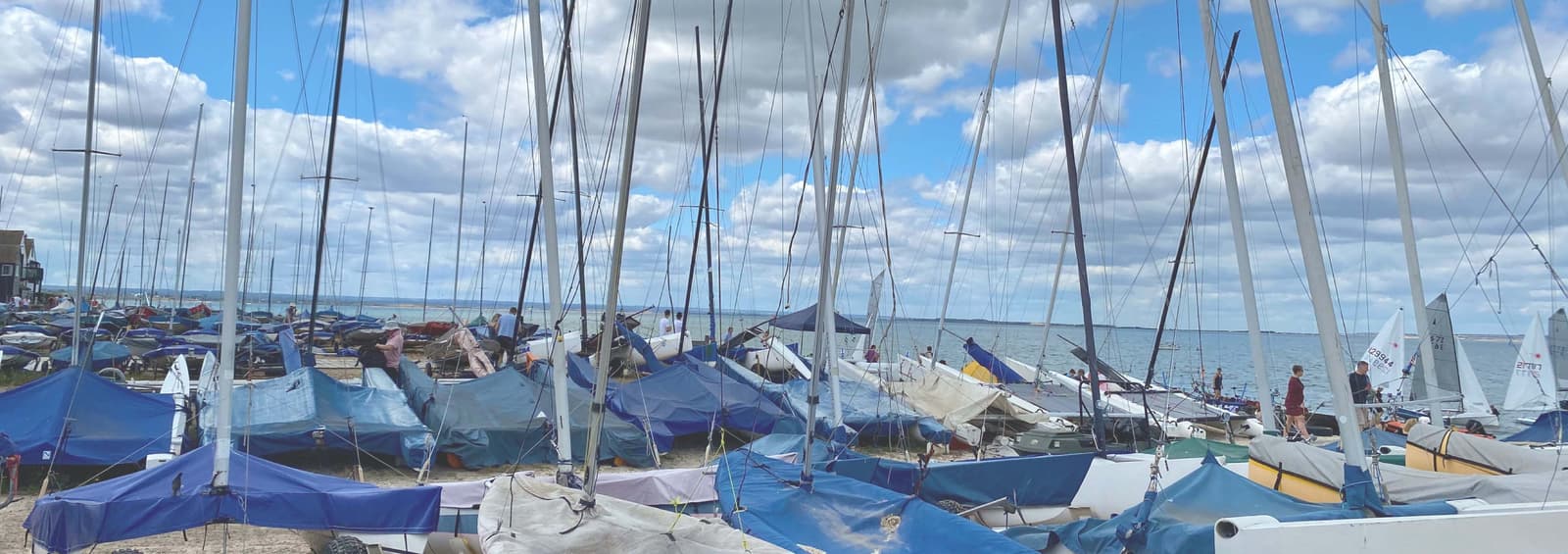 A collection of sailboats with colorful tarps are parked on a sandy shore under a partly cloudy sky. Tall masts reach upwards, while the calm sea and distant horizon can be seen in the background.
