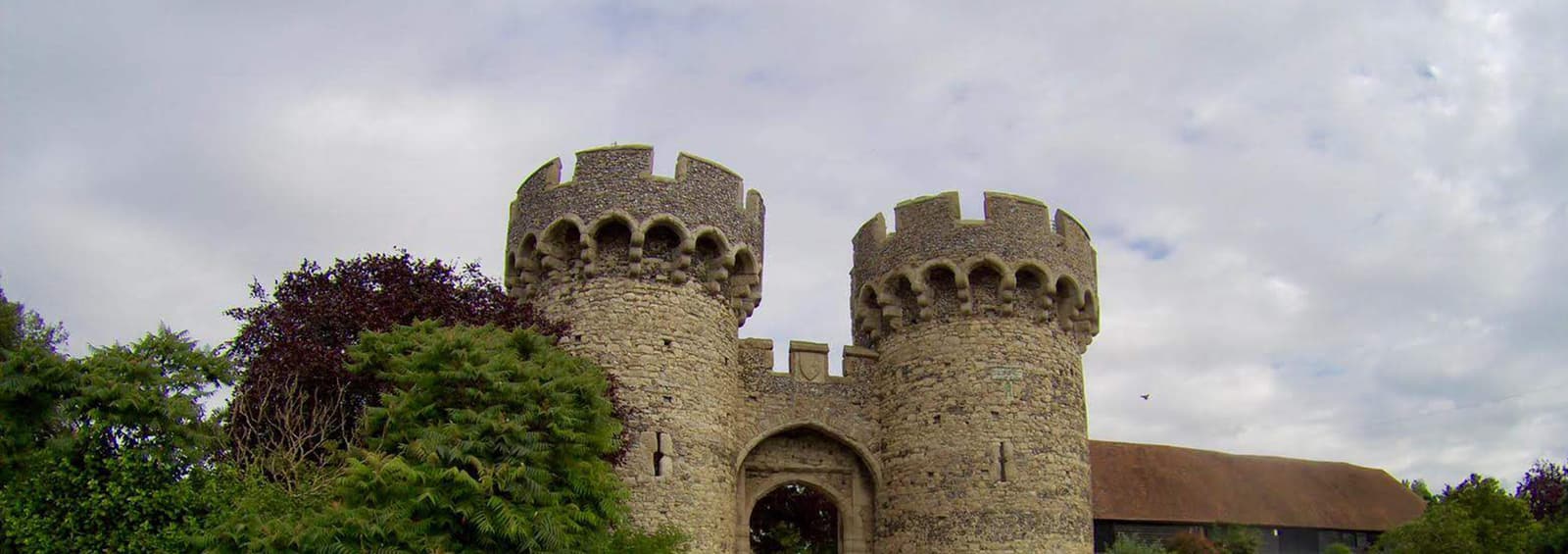 A historic stone gatehouse with two round towers, surrounded by lush greenery and under a cloudy sky. The towers feature crenellations and an arched entrance at the center. A building with a sloped roof is partially visible on the right.