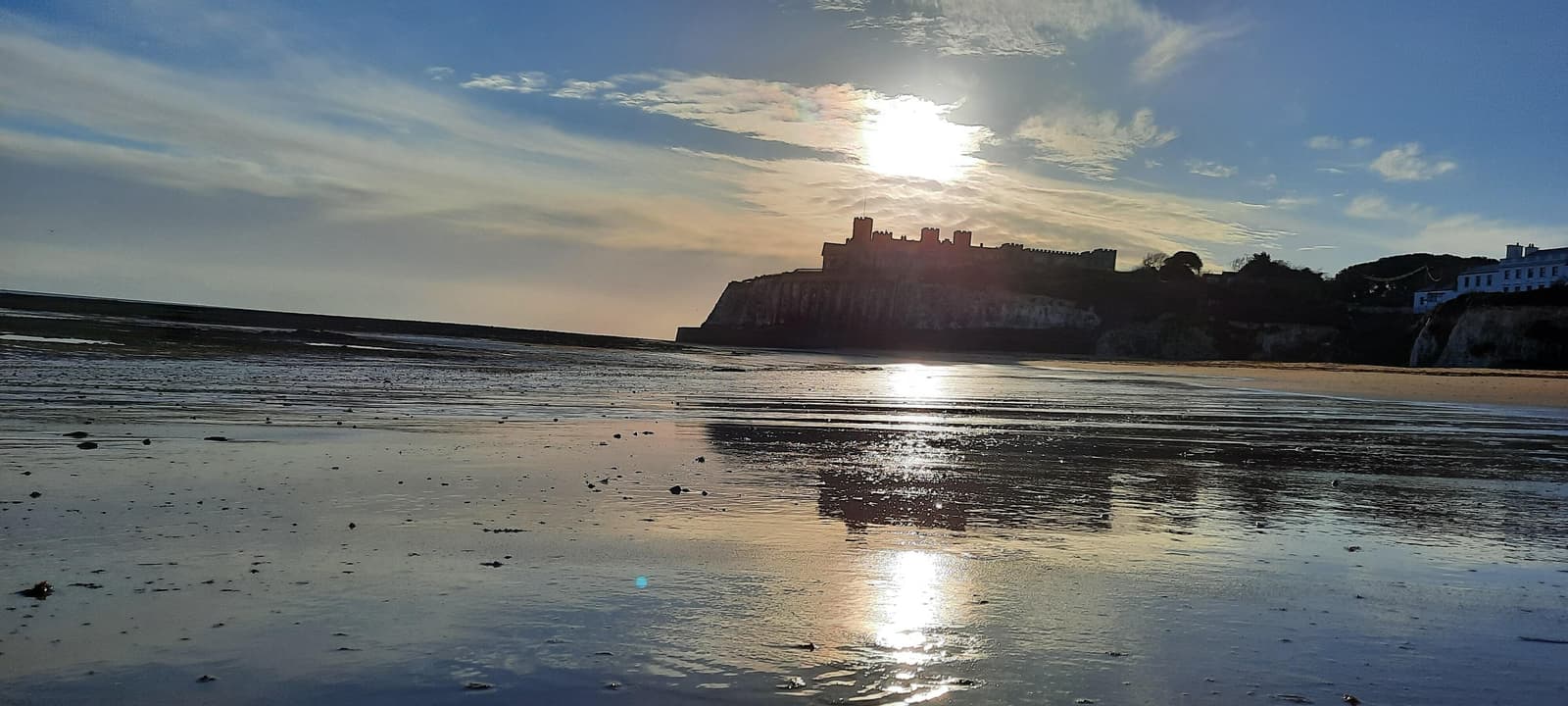 Sunset over a coastal landscape with a cliffside silhouette of a castle. The sandy beach reflects the sky's colors, while gentle waves lap the shore. Clouds disperse the sunlight, creating a serene and picturesque scene.
