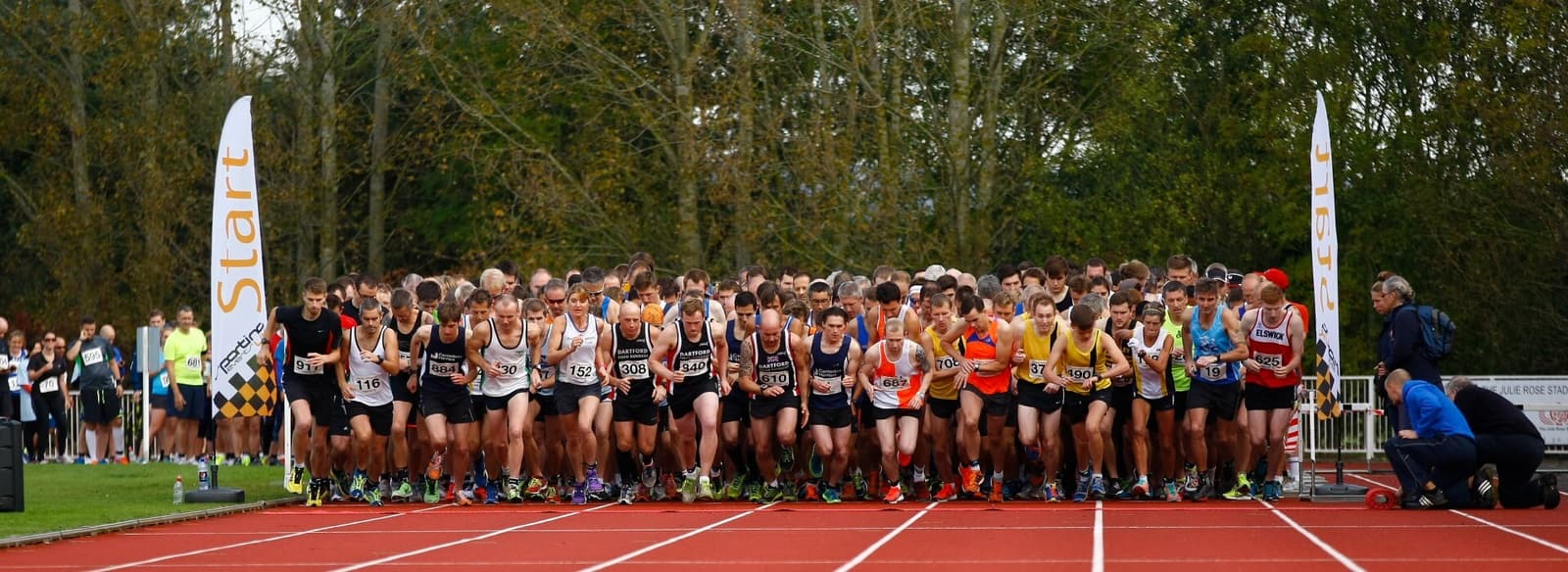 A large group of runners at the starting line of a race on a track. They wear numbered bibs and varied athletic gear. Bright banners reading "Start" are visible, and trees are in the background.