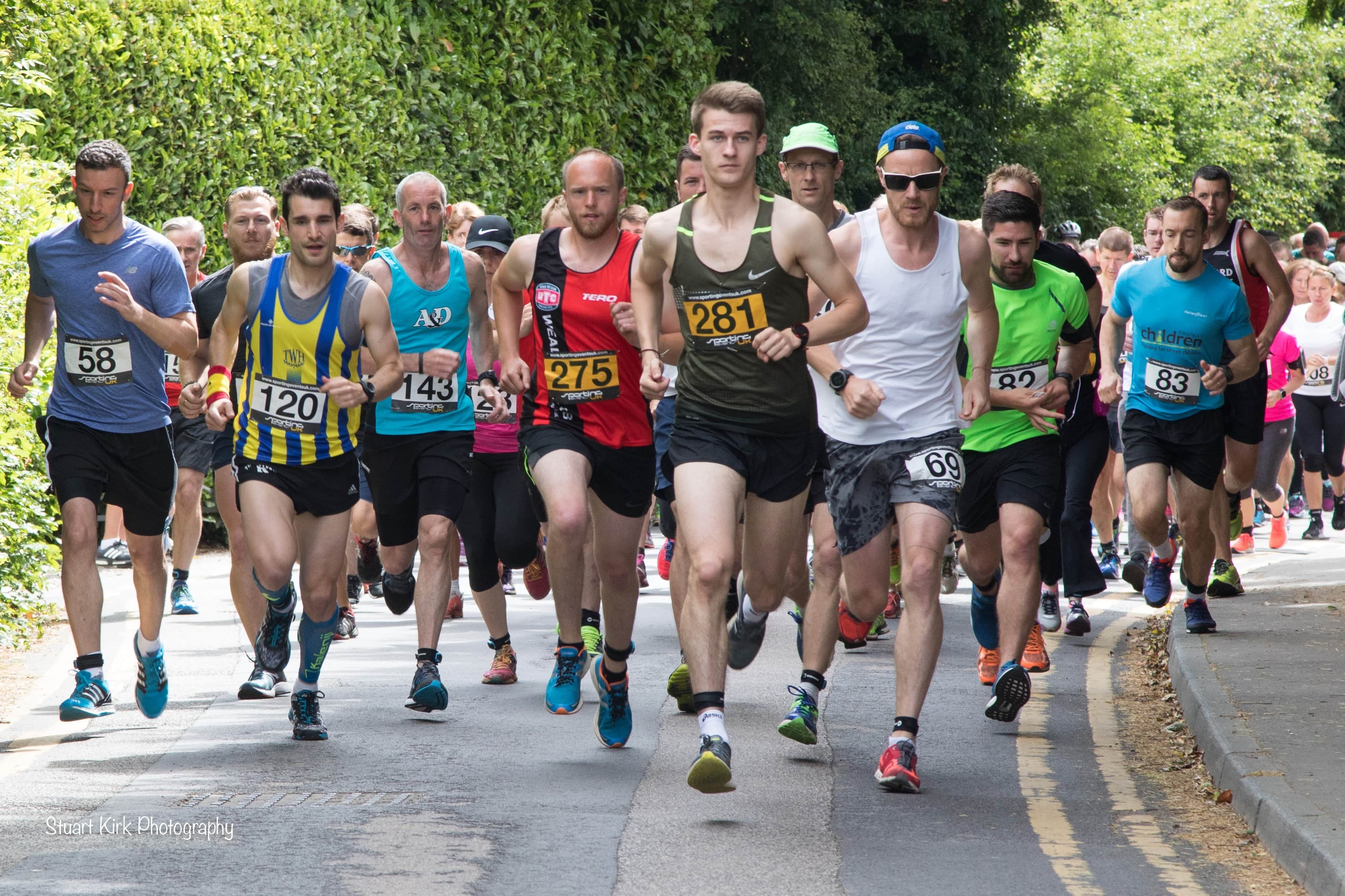 A group of runners, wearing numbered bibs, start a race on a tree-lined street. The participants are dressed in various athletic wear, and the atmosphere appears energetic and competitive.
