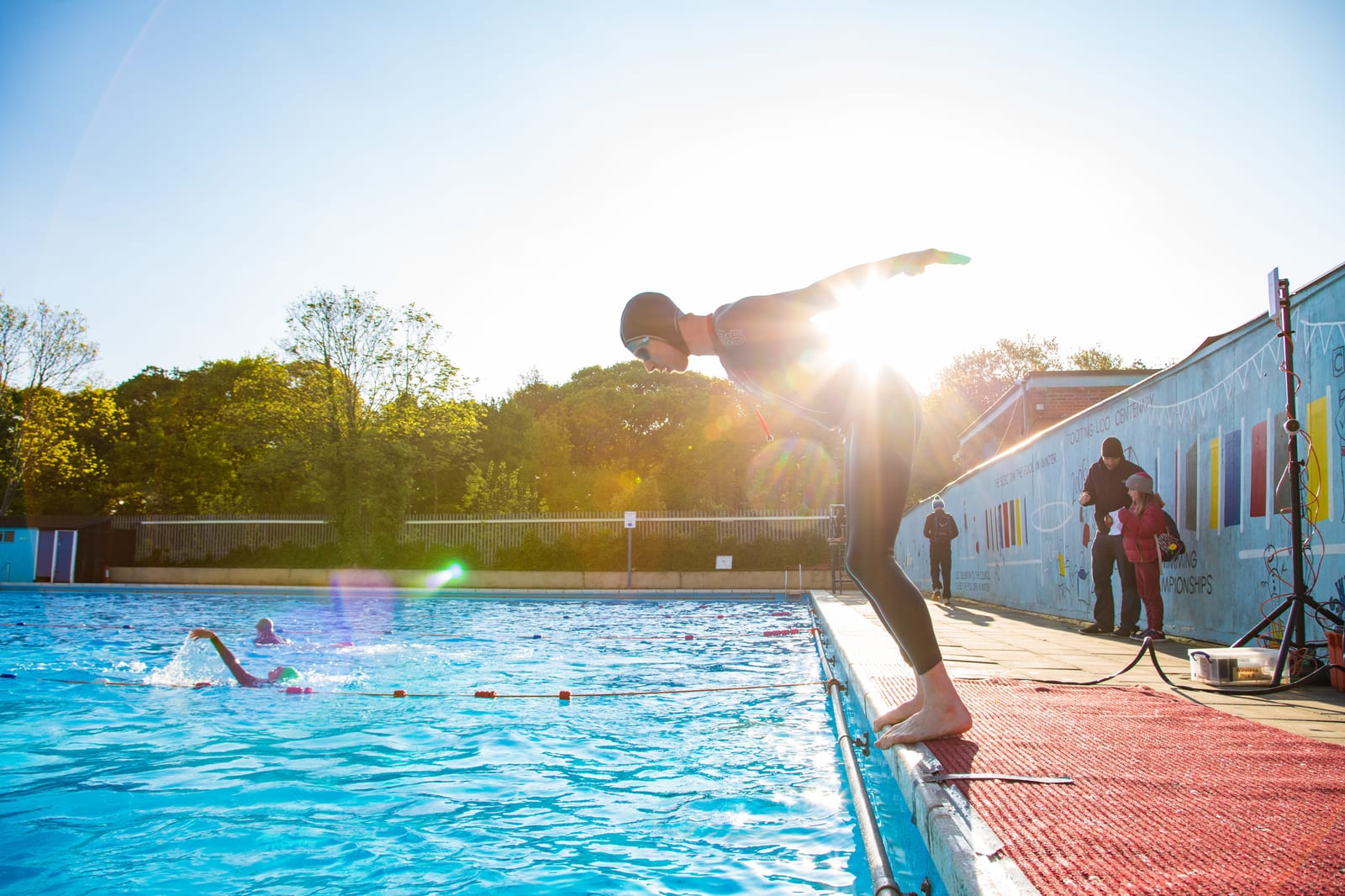 A swimmer in a wetsuit and cap stands poised to dive into a pool at the edge, with the sun shining brightly behind them. Another swimmer can be seen in the background, swimming laps. The poolside has a few people and equipment set up. Trees line the background.
