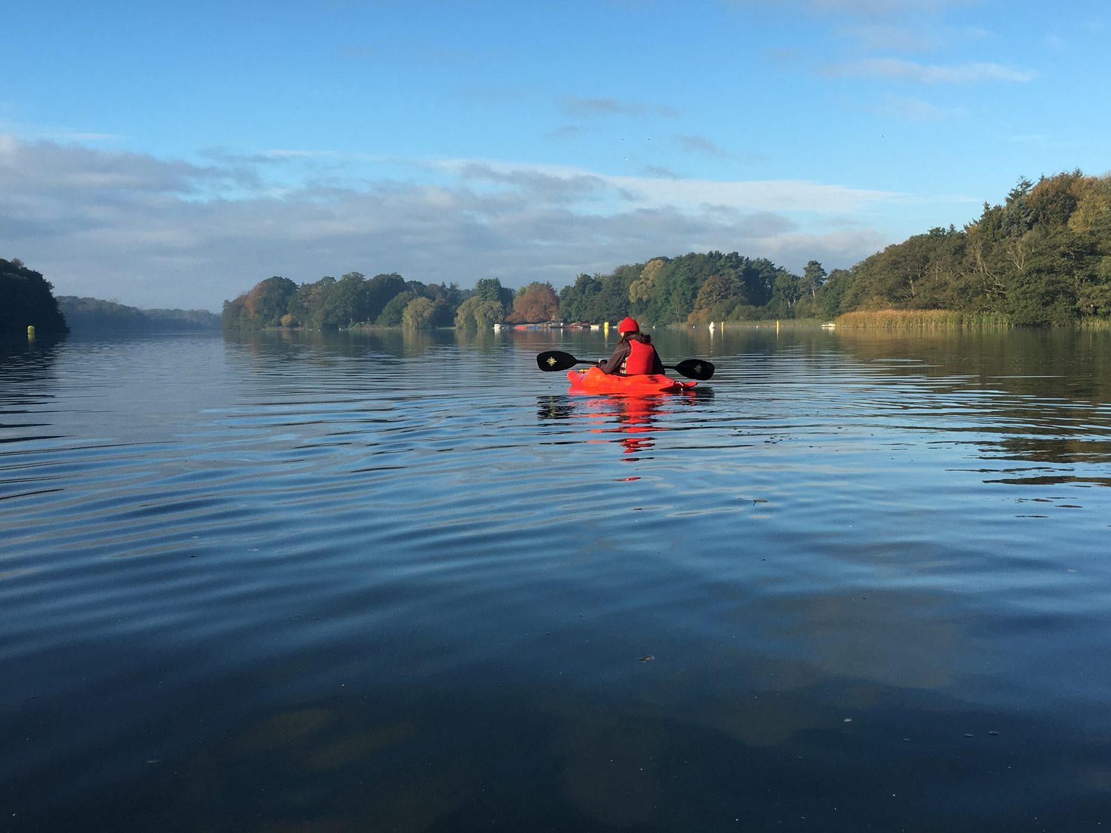 A person in a red kayak paddles across a calm lake surrounded by trees under a blue sky with scattered clouds. The water reflects the trees and sky, creating a serene and peaceful scene.