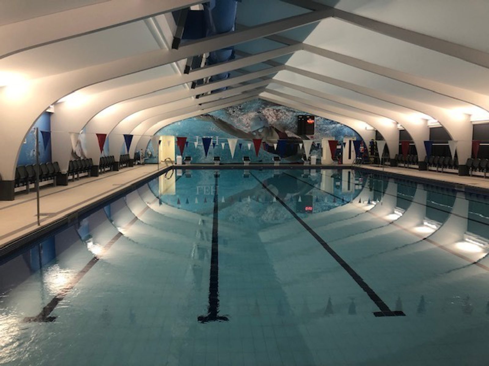A well-lit indoor swimming pool with clear water and marked lanes. The pool is surrounded by white arched beams and has colorful triangular flags hanging above the lanes. The walls feature aquatic-themed murals, and there are rows of black chairs on both sides.