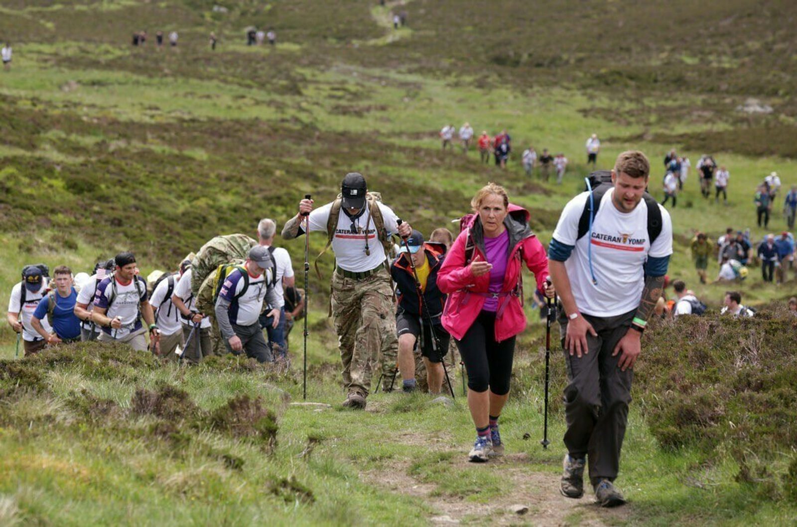 A large group of hikers climbs a grassy, slightly rocky hillside. They are wearing outdoor gear, including backpacks and hiking poles. The landscape is lush and green, with a mix of open grass and patches of shrubs. The sky appears to be overcast.