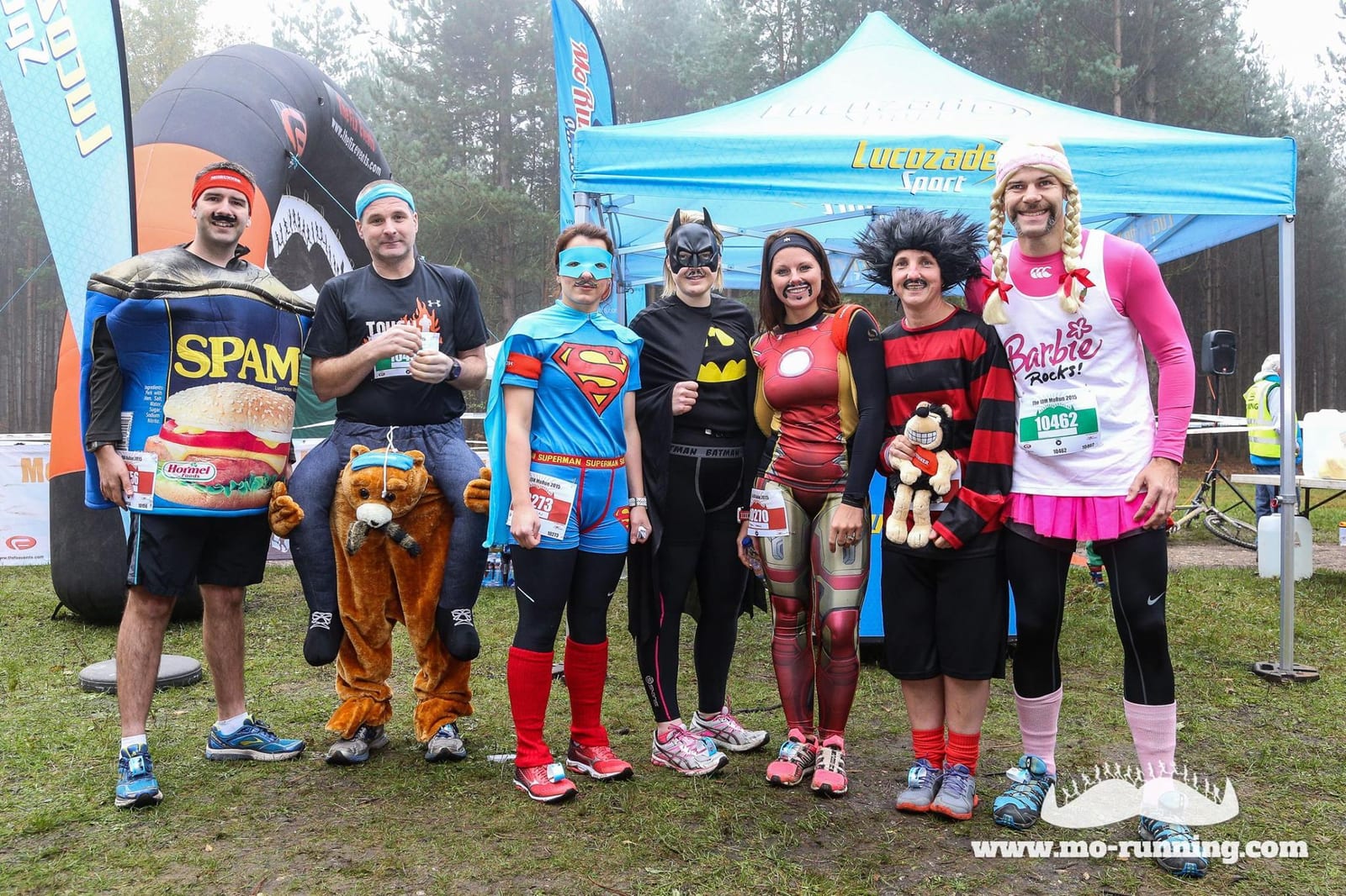 A group of people dressed in various costumes, including a can of Spam, a cowboy riding a horse, Superman, Batman, Iron Man, a clown-like figure, and Barbie. They are standing in front of a blue tent at an outdoor event.
