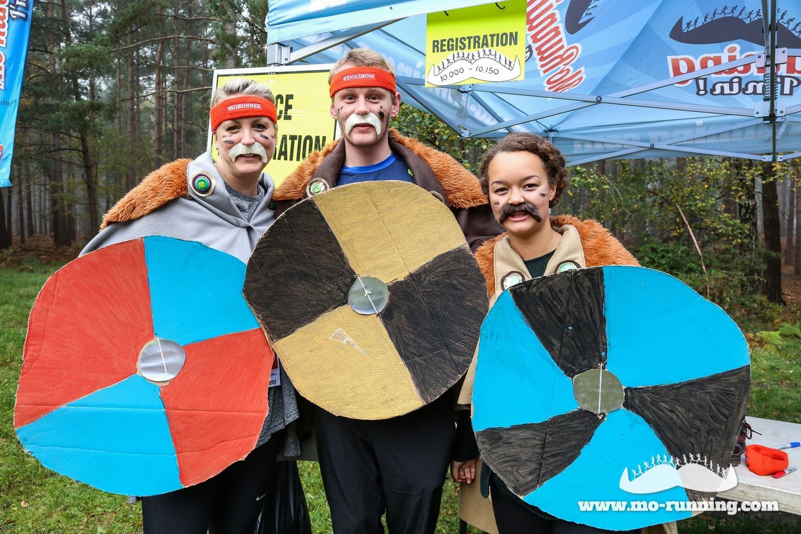 Three people dressed in costumes in front of a registration booth. They wear fur capes, headbands, and fake mustaches, and hold large, round, painted shields. The background includes a forest and banners for a "Mo-Running" event.