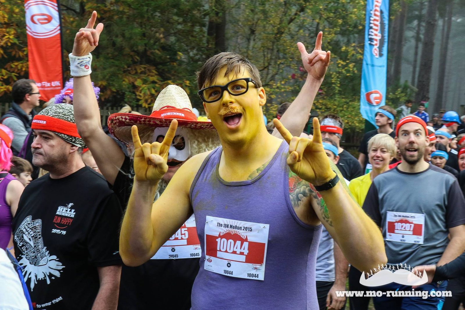 A man dressed as a yellow cartoon character with glasses and wearing a tank top shows rock-and-roll hand signs at a running event. He stands among other runners, some in costumes, in an outdoor setting. Visible race numbers include "10044". A website URL is visible.