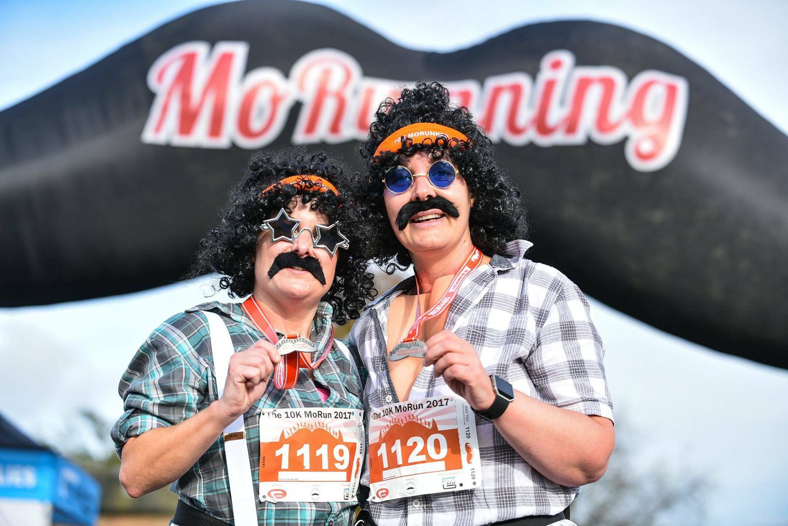 Two people dressed in matching wigs, fake mustaches, and sunglasses hold medals while posing in front of an inflatable mustache. They both have race bibs with the numbers 1119 and 1120. The event banner behind them reads "MoRunning.