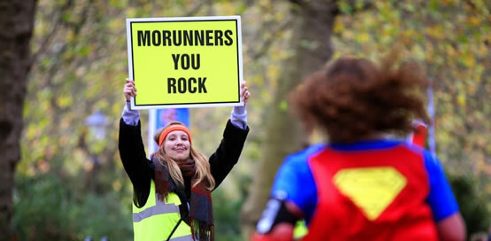 A person stands outdoors wearing a neon safety vest and holding a large yellow sign that reads "MORUNNERS YOU ROCK." They are cheering as runners, some wearing colorful costumes, participate in a race. Trees in the background suggest the event is taking place in a park.