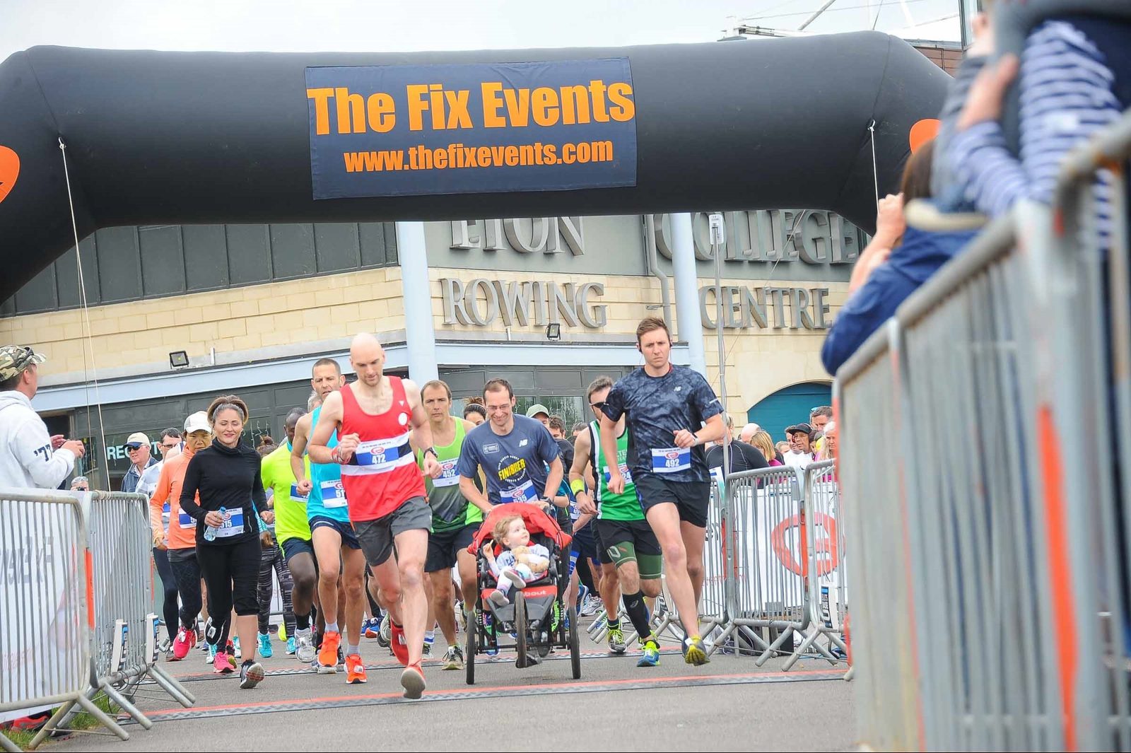 A group of runners begin a race, passing under an inflatable arch with "The Fix Events" written on it. One runner is pushing a person in a wheelchair. Spectators stand by at the sides, some taking photos. The background shows a building with signage for a rowing center.