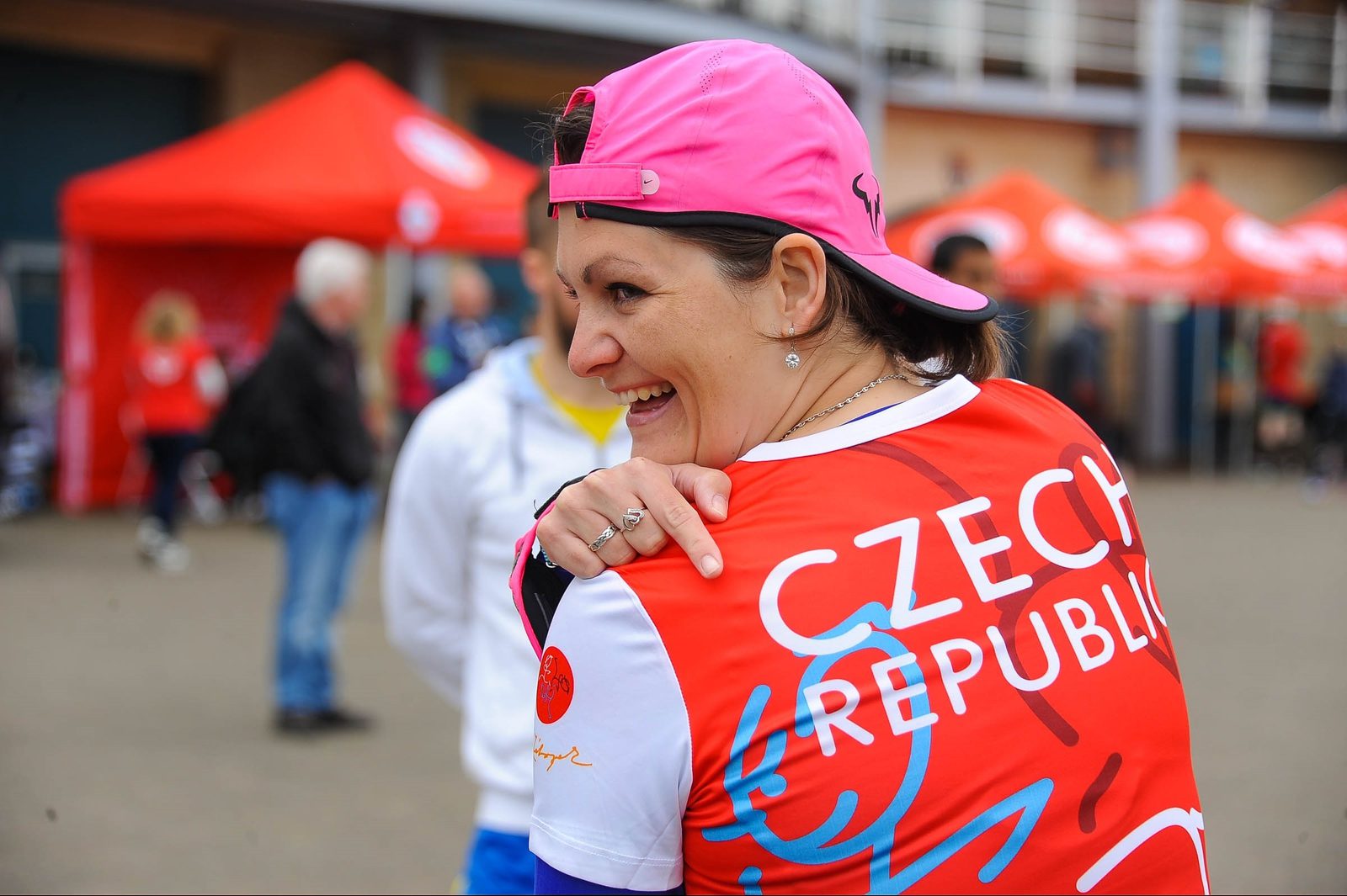 A woman wearing a red and white "Czech Republic" shirt and a pink cap smiles brightly. She is standing outside with several people, red tents, and a building in the background.