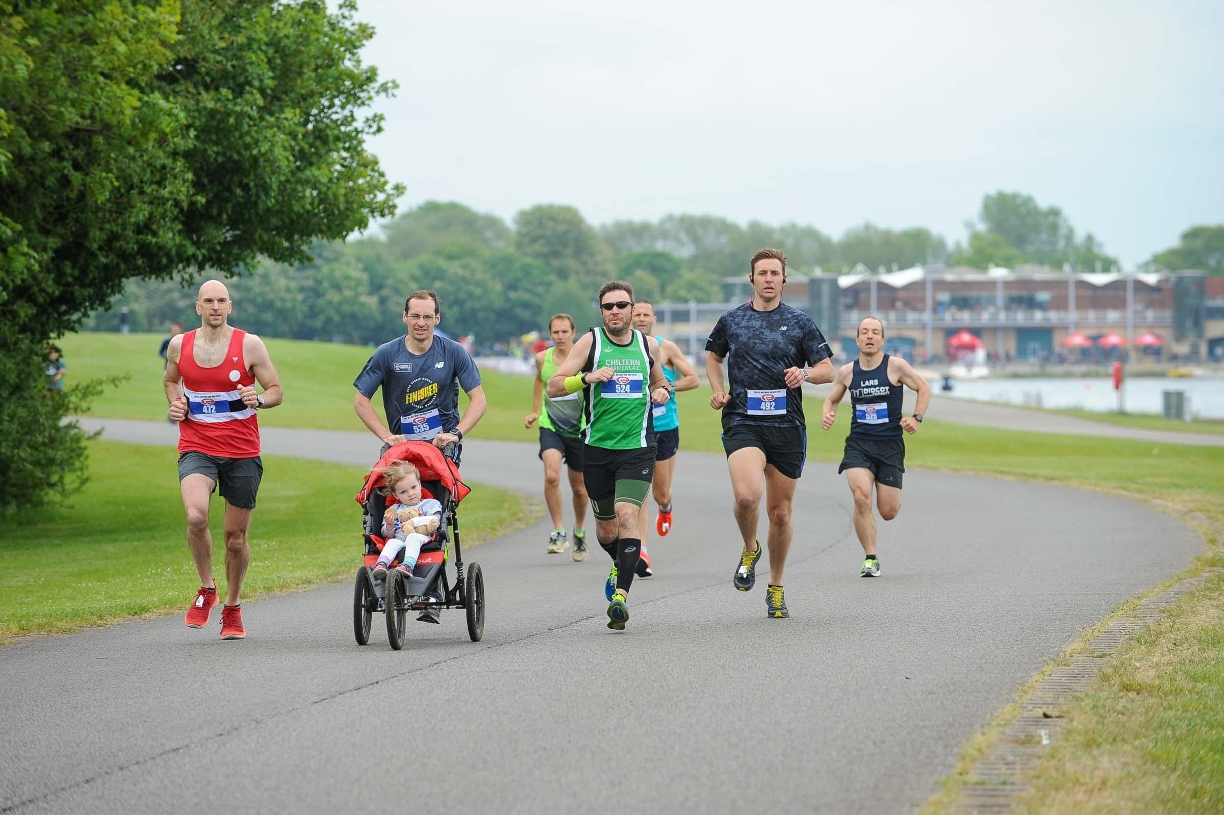 A group of runners is participating in a race on a paved path next to a body of water. One runner is pushing a child in a stroller. Trees and park facilities are visible in the background, and the weather appears clear and pleasant.