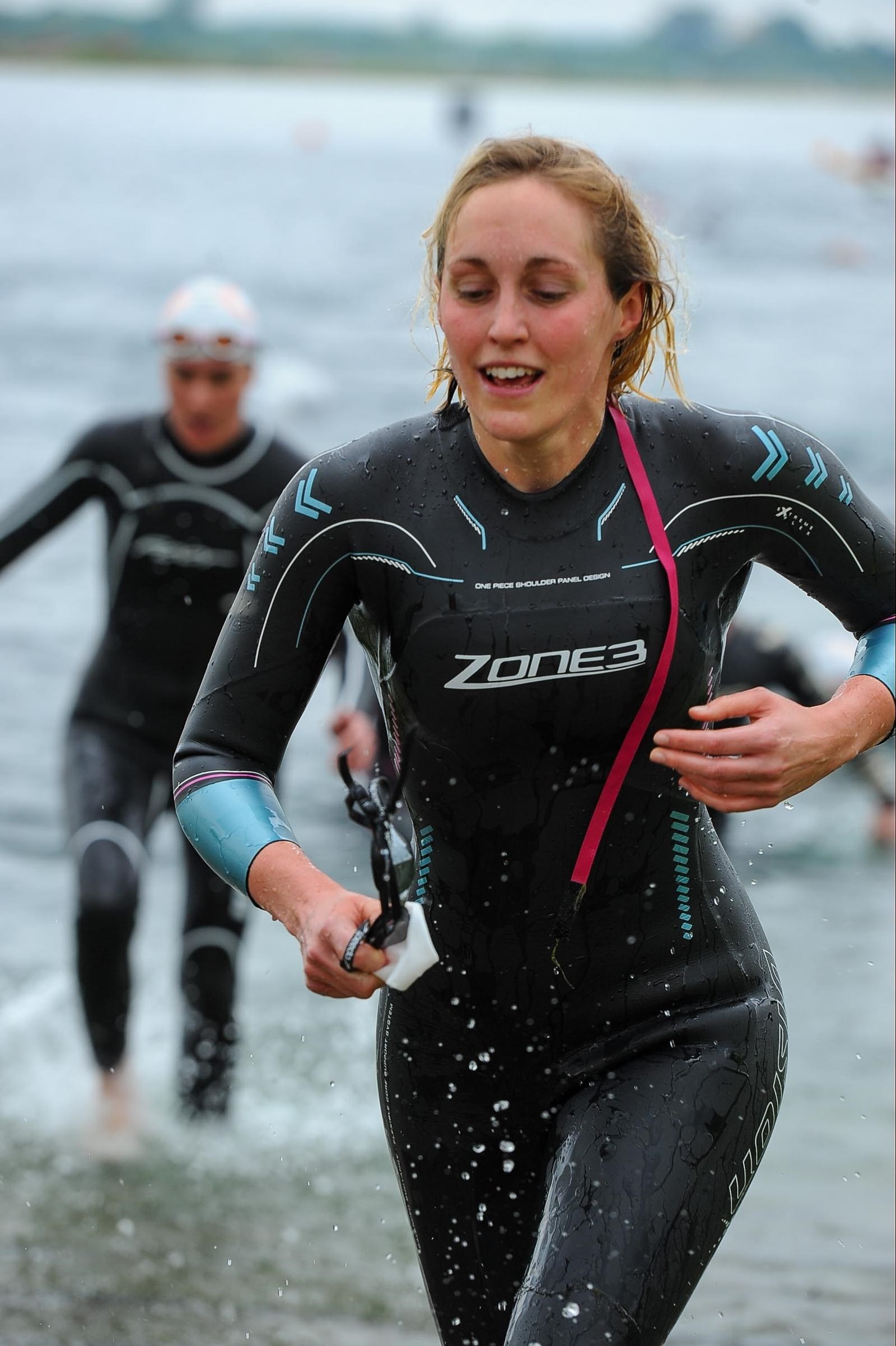 A female triathlete emerges from a body of water, smiling and breathing heavily, with goggles in hand. She is wearing a wetsuit with the brand name "Zone3" visible. Another athlete is seen in the background, also wearing a wetsuit and goggles, nearing the shore.