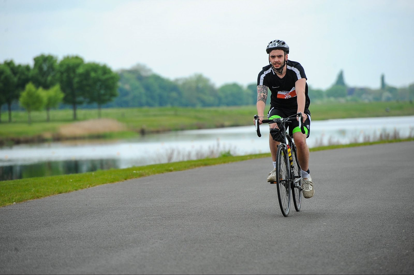 A man wearing a helmet and sportswear rides a bicycle on a paved path beside a body of water. Trees and grassy areas are in the background. The weather appears clear and calm.