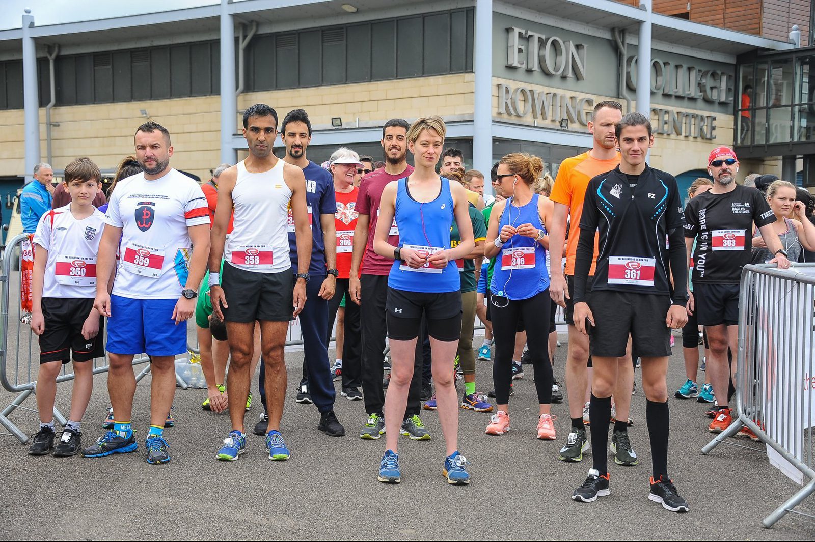 A group of runners stands at the starting line of a race outside of Eton College Rowing Centre. They wear various athletic outfits, some with race bibs. The atmosphere is anticipatory as they await the start signal.