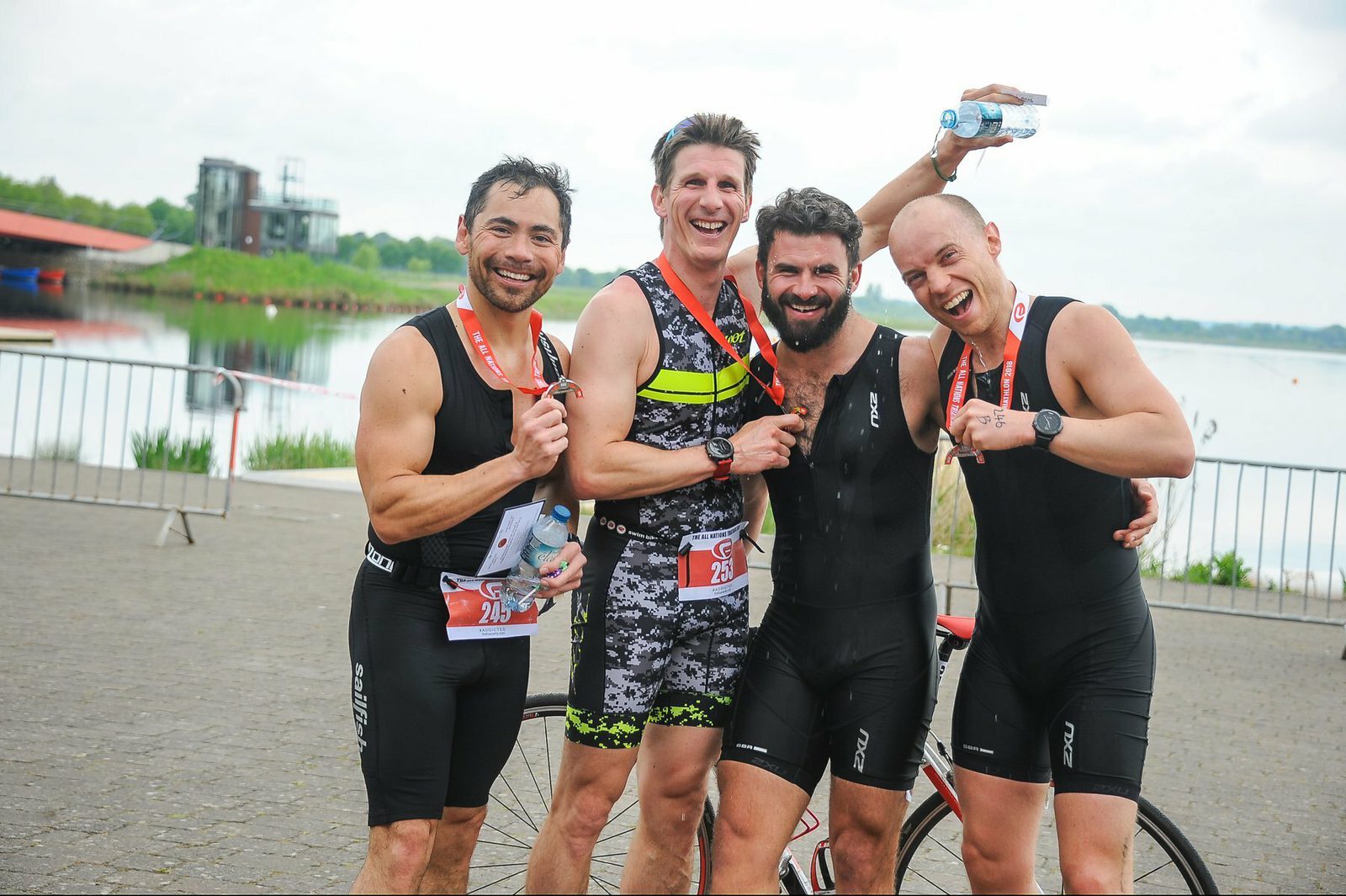 Four men wearing athletic gear stand together, smiling and holding race medals around their necks. They are positioned in front of a lake, with some holding water bottles and a race bib numbered 26 visible. The atmosphere is celebratory and joyful.