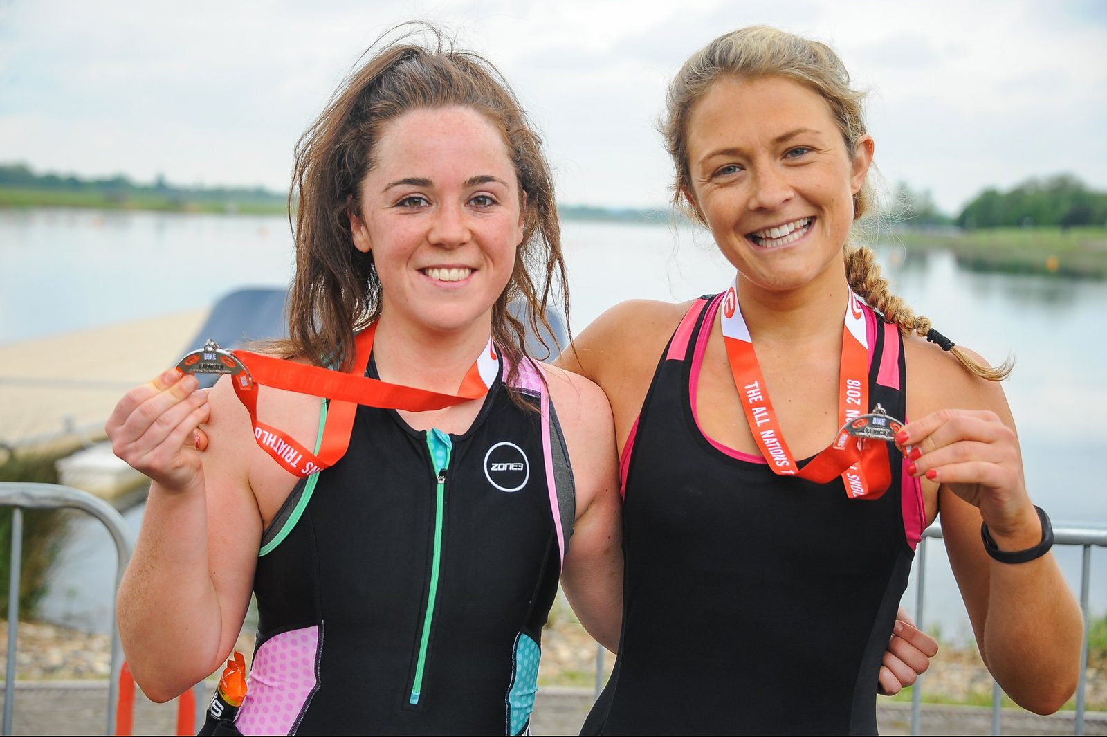 Two women in athletic gear stand side by side, smiling and displaying medals around their necks. The background features a body of water and a dock. Both women wear black athletic suits, and their medals hang from red lanyards. They appear happy and accomplished.