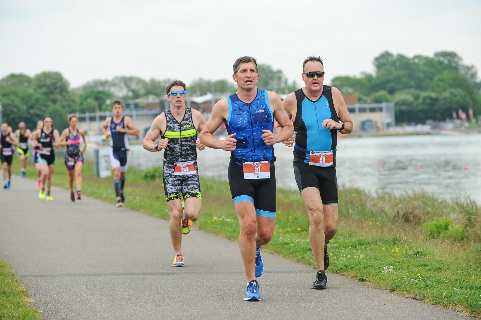 A group of athletes participating in a triathlon are running on a paved path adjacent to a body of water. The men in the foreground wear blue and black athletic gear with race numbers. Green trees and buildings can be seen in the background under a cloudy sky.