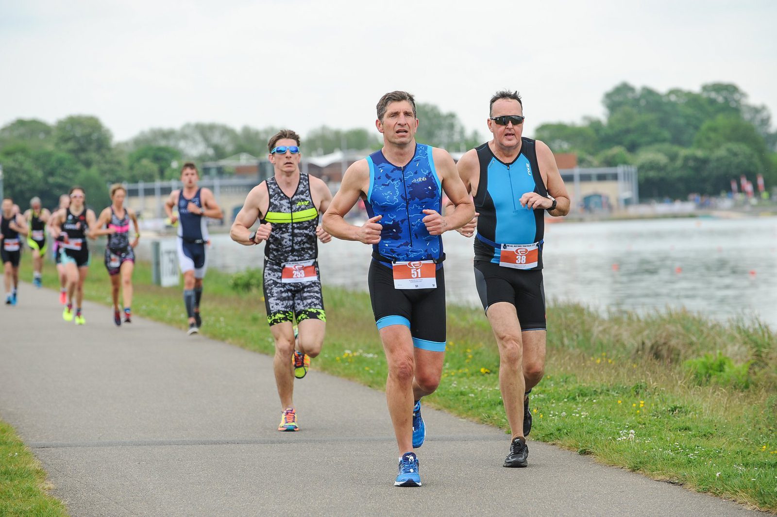 A group of athletes running along a paved path next to a water body during a race. They wear race bibs and athletic gear; most have numbers displayed prominently on their shorts. The background features trees and a building, indicating an outdoor event.