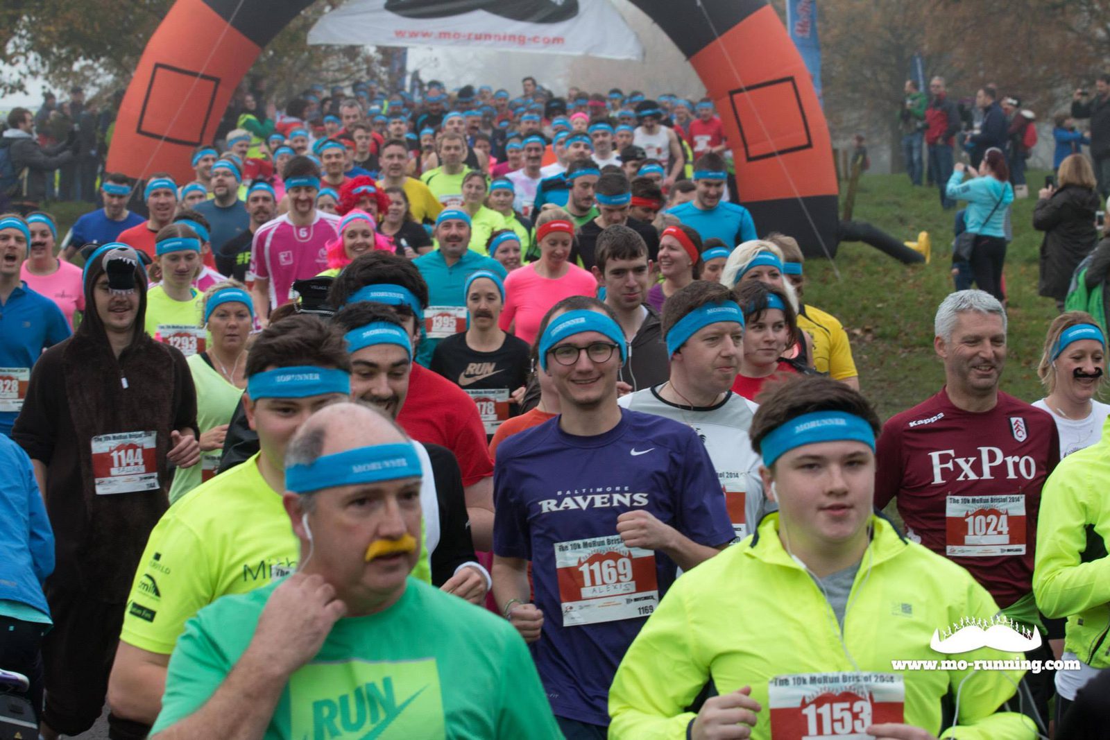 A large group of runners, many wearing blue headbands and colorful athletic gear, participate in a race. Several people sport comedic fake mustaches. An inflatable arch marking the race's start is visible in the background. The mood seems lively and energetic.