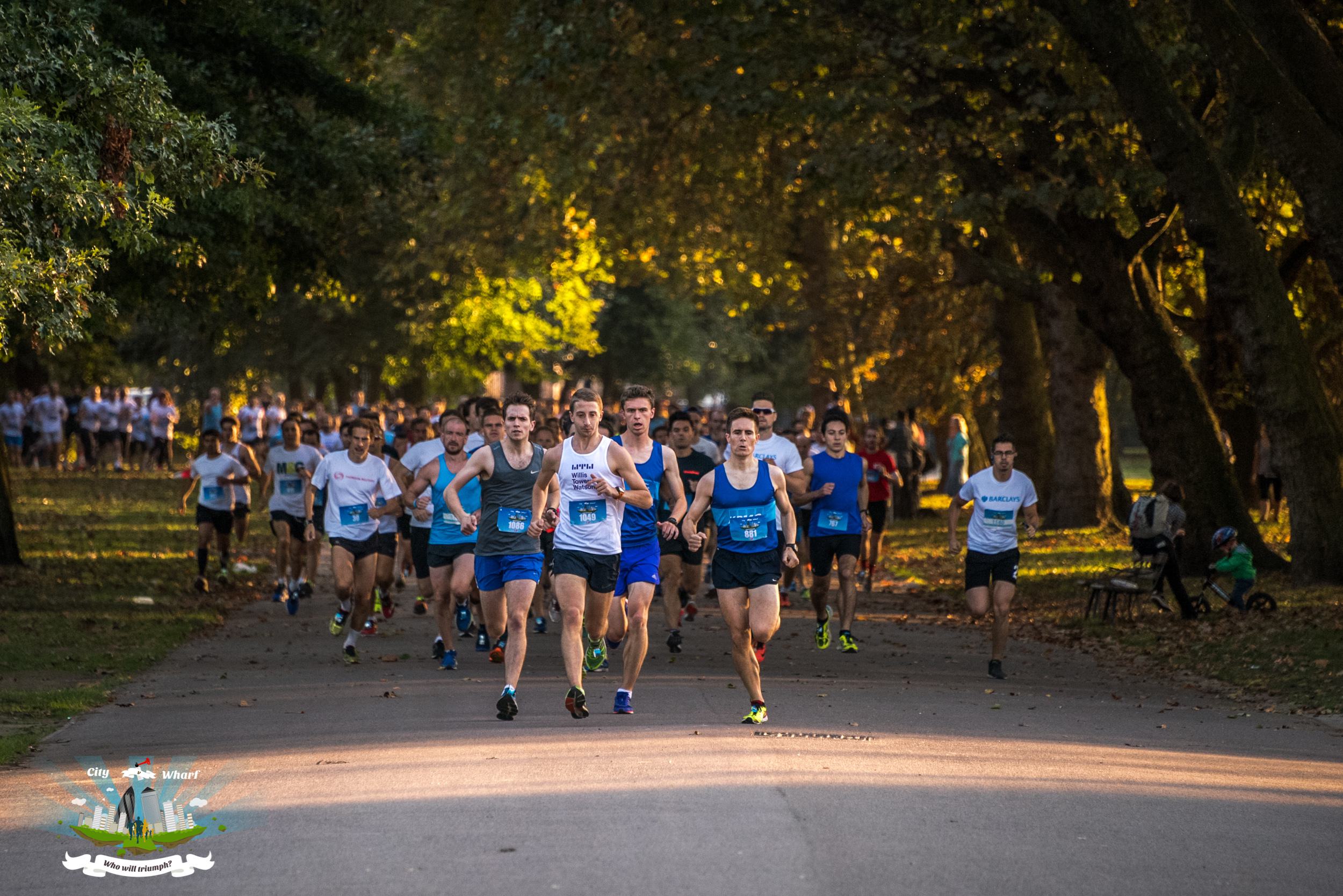 A group of runners in athletic gear participate in a race on a tree-lined path in a park. The sun casts a warm glow, highlighting the runners and greenery. Spectators are visible in the background.
