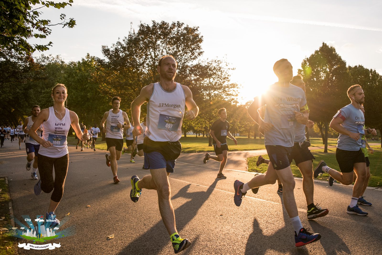 A group of runners participating in a race on a paved path in a park during sunset. They are all dressed in athletic gear, with race bibs on their shirts. Sunlight filters through the trees in the background, creating a warm glow over the scene.