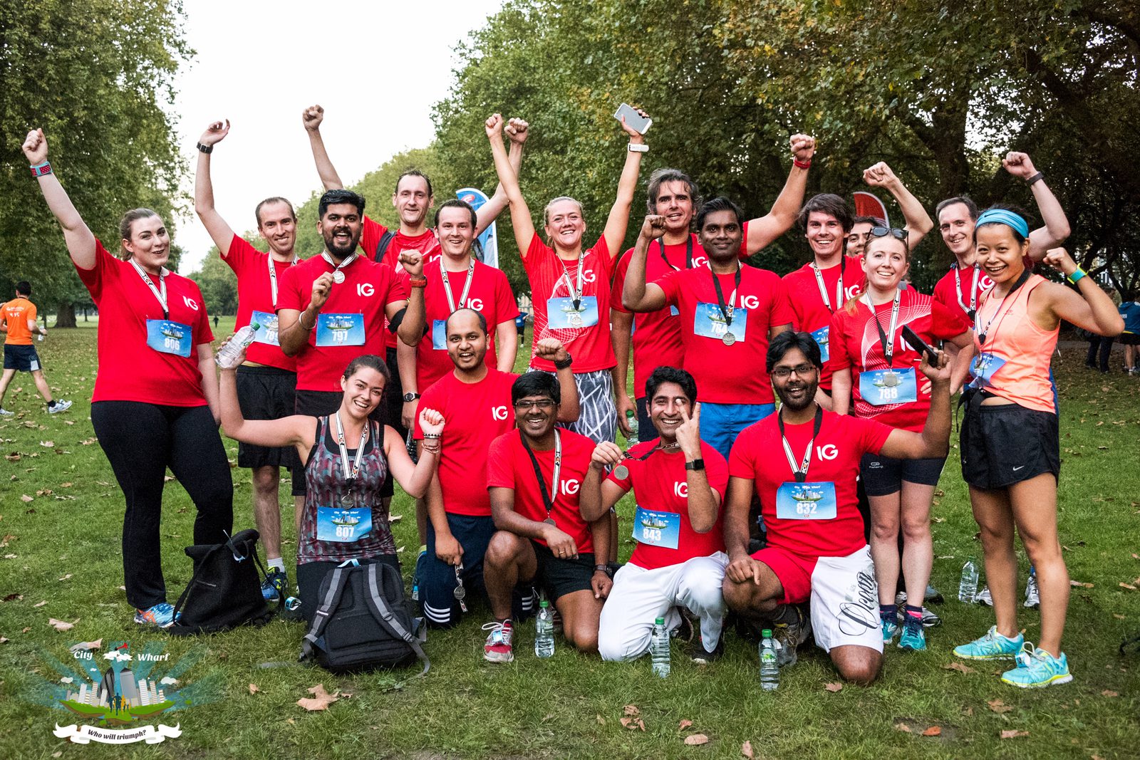 A diverse group of people wearing red shirts and medals pose triumphantly with raised arms in a park. They are gathered closely together, smiling and celebrating their achievement. Some are kneeling in the front while others stand behind, surrounded by greenery.