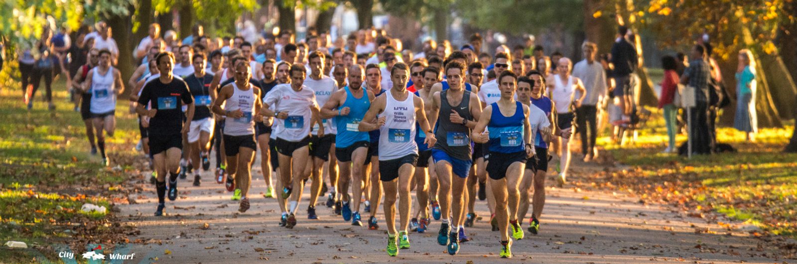 A large group of runners in athletic gear participating in a race in a park setting, surrounded by trees with green and autumn leaves. Some onlookers and supporters are visible in the background, cheering the runners on. The sky appears clear and the weather is sunny.