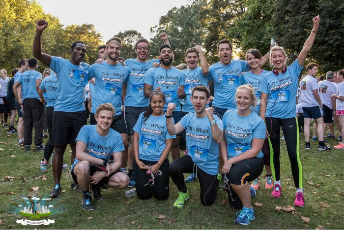 A group of people wearing matching blue "City Wharf" t-shirts pose together outdoors on grass. Some stand with their fists raised, while others kneel or sit in front. They appear happy and energetic, possibly participating in a team event or charity run. Trees are in the background.