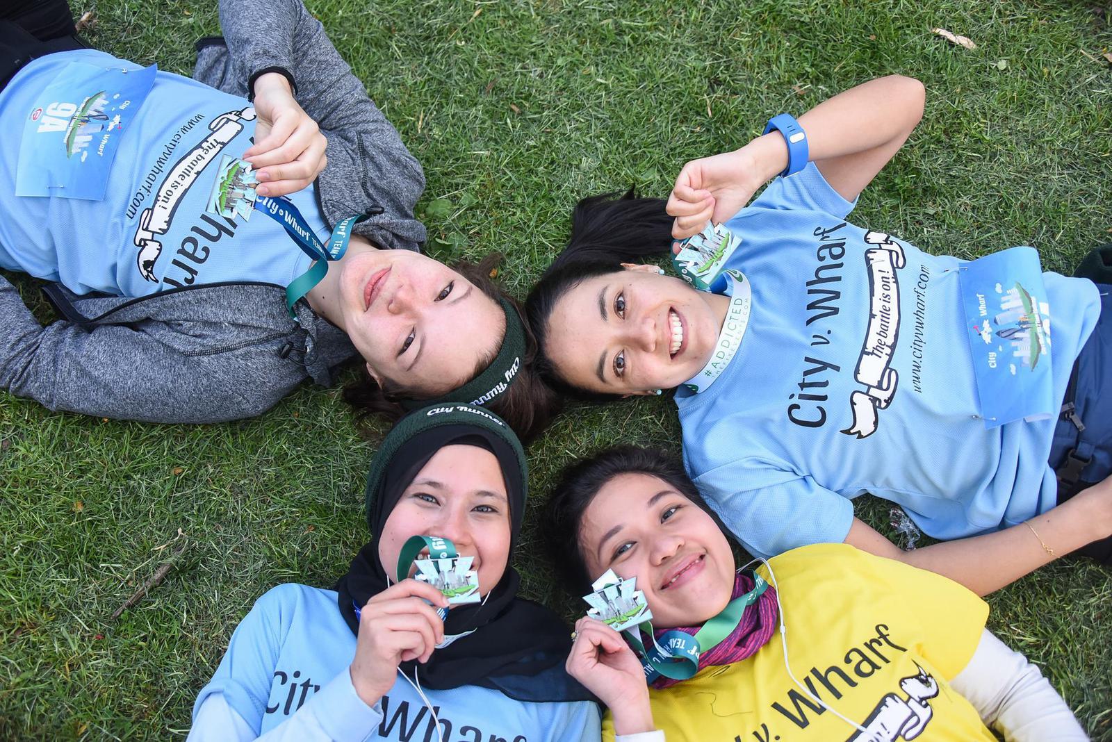 Four people lie on grass, wearing medals and colorful shirts. They smile at the camera, each holding their medal. The shirts read "City v. Wharf," and they appear to be celebrating after a race or event.