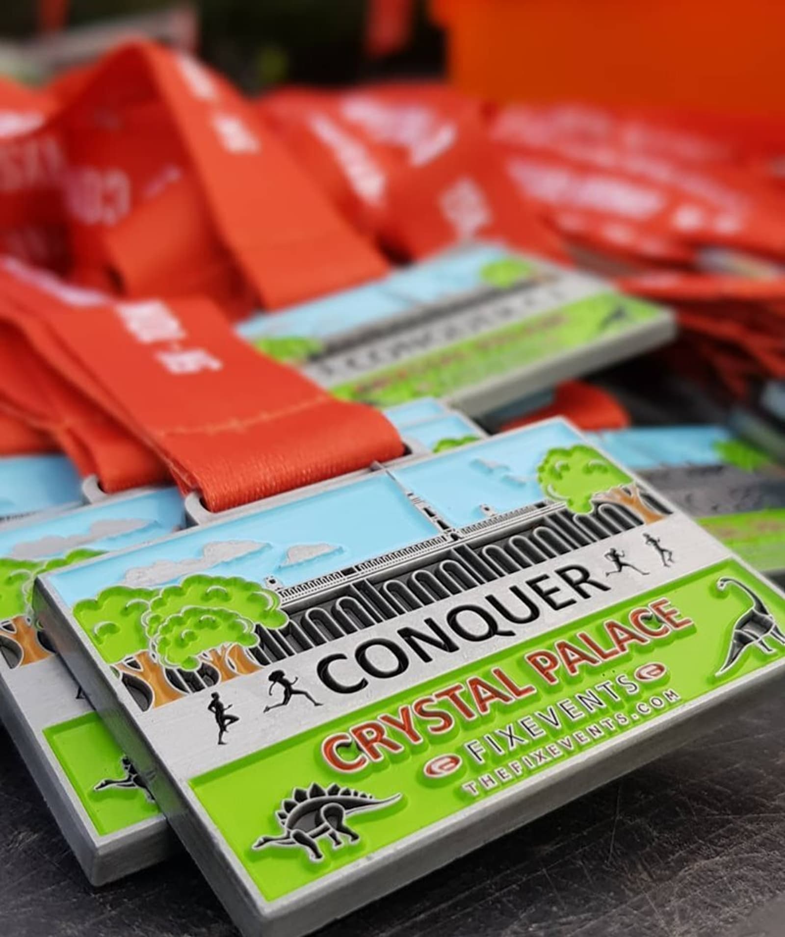 A stack of colorful "Conquer Crystal Palace" medals with red ribbons. The medals feature an image of a building, trees, runners, and a dinosaur, along with text reading "Conquer," "Crystal Palace," and "Fix Events" with their website.