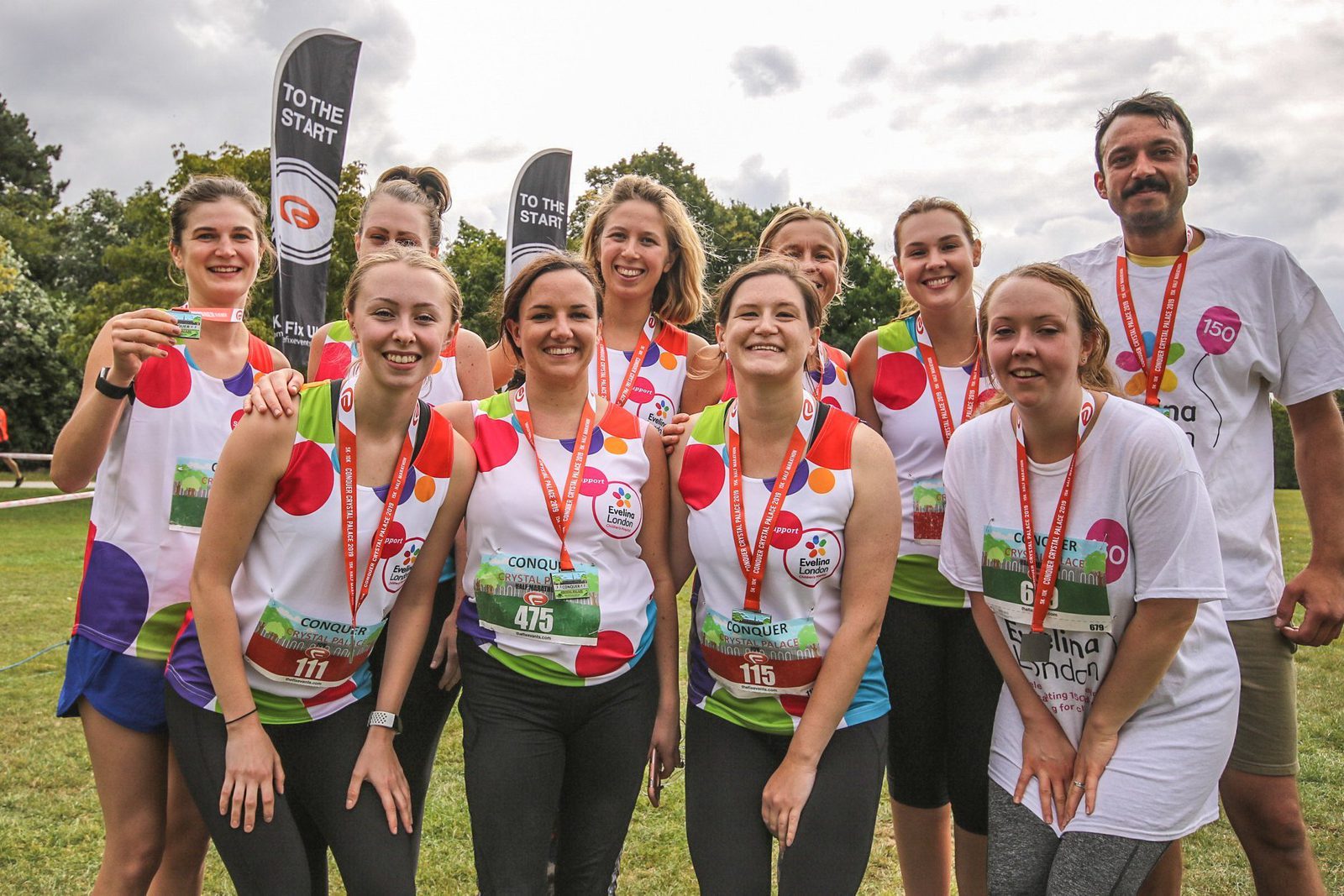 A group of ten smiling people, all wearing white T-shirts with colorful polka dots and red medals around their necks, pose together outdoors on a grassy area. Some people hold up their medals proudly. Two banners in the background read "TO THE START.