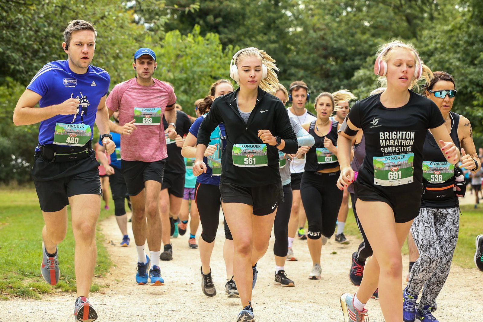 A group of runners participating in a marathon. They are running outdoors on a gravel path surrounded by trees. Many contestants are wearing race bibs and headphones, focused on the race. The atmosphere looks lively and competitive.
