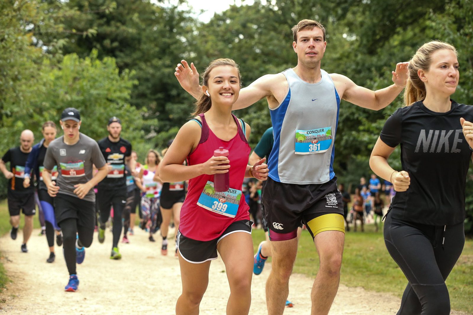 A group of runners, men and women, participating in an outdoor race on a dirt trail surrounded by trees. Two runners in the center, a woman in a red tank top and a man in a blue tank top, are smiling while running. Other participants are visible in the background.