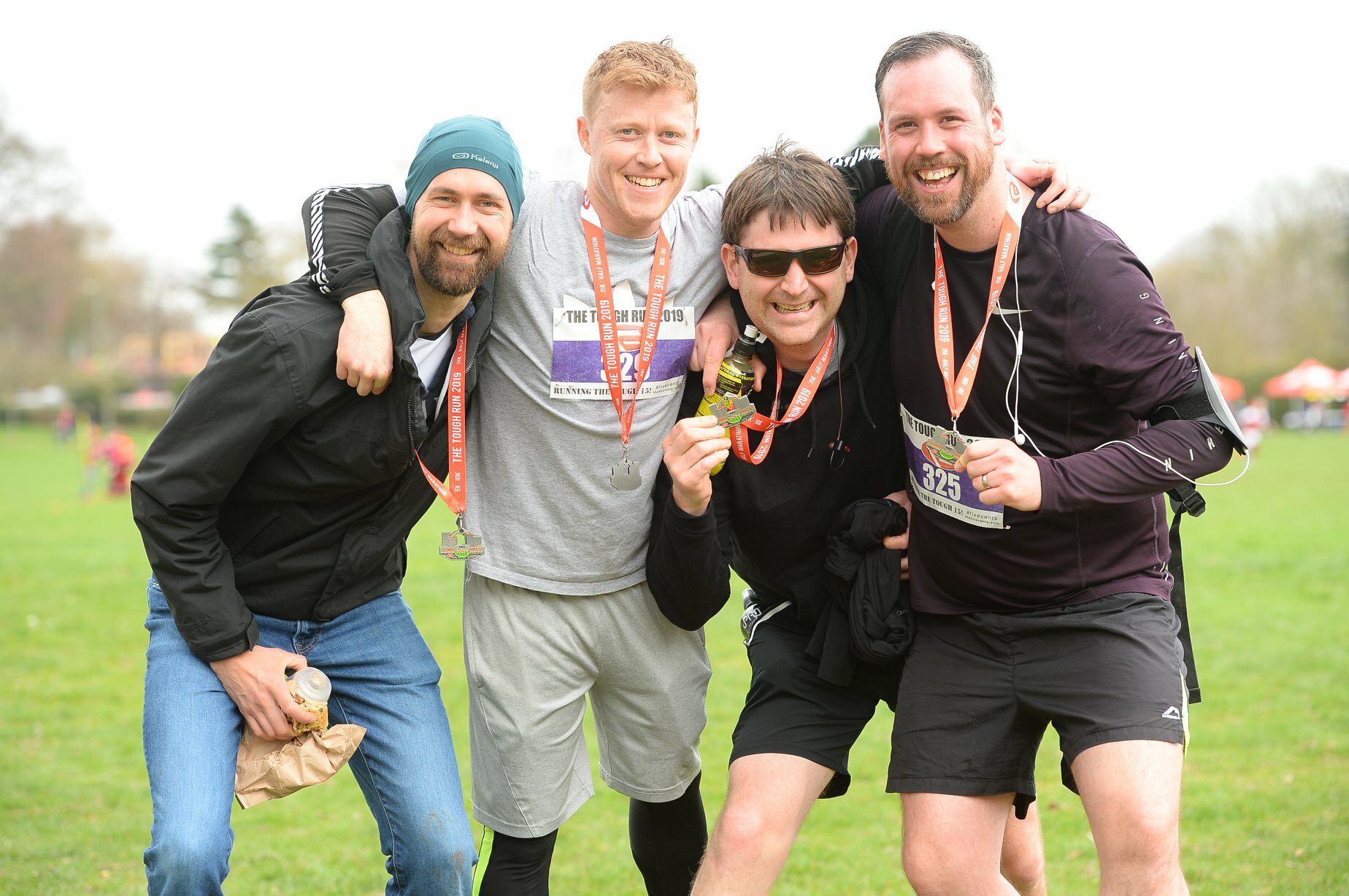 Four smiling men pose together outdoors after a race, each wearing a finisher's medal. They are dressed in athletic gear, with some sporting sunglasses and beanies. The background shows a grassy field and other participants.