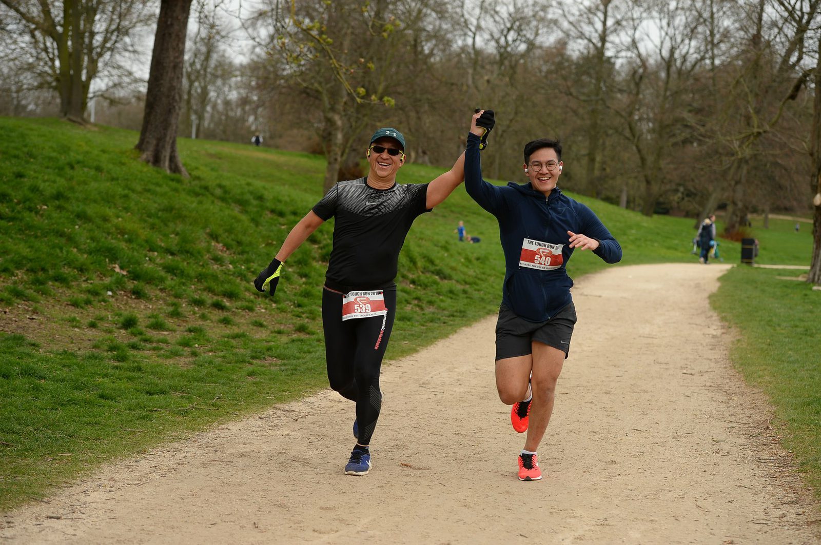 Two male runners in athletic gear run on a dirt path through a park, holding hands and smiling triumphantly. Both wear race bibs, one in black attire and the other in a dark blue outfit. People and trees are visible in the background.