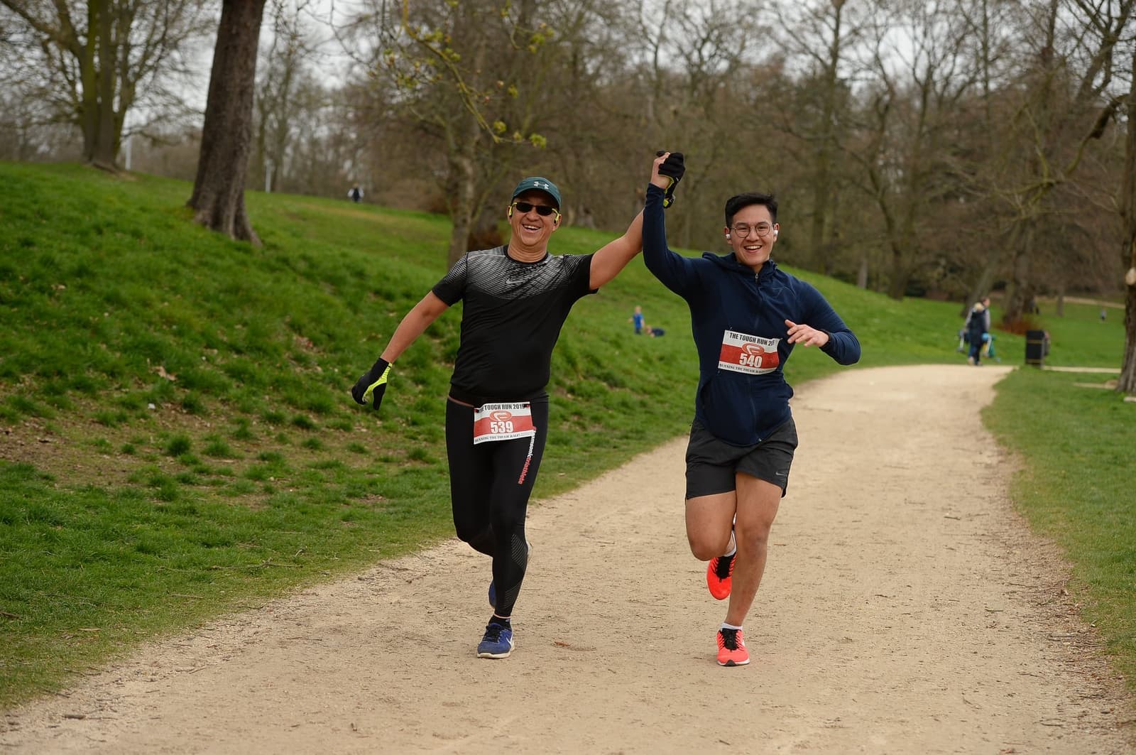 Two men, dressed in athletic gear with race bibs, run side by side on a park trail, holding hands in celebration. They are smiling and appear jubilant, surrounded by green grass and trees. Other people are seen in the background.
