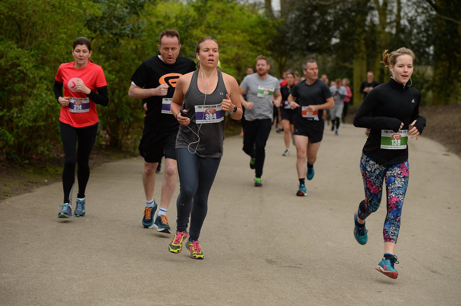 People running in a park during a race, wearing various athletic outfits and race bibs. The path is surrounded by green foliage, and the runners display a range of expressions, focusing on the task. It's a cloudy day, evident from the diffused lighting.