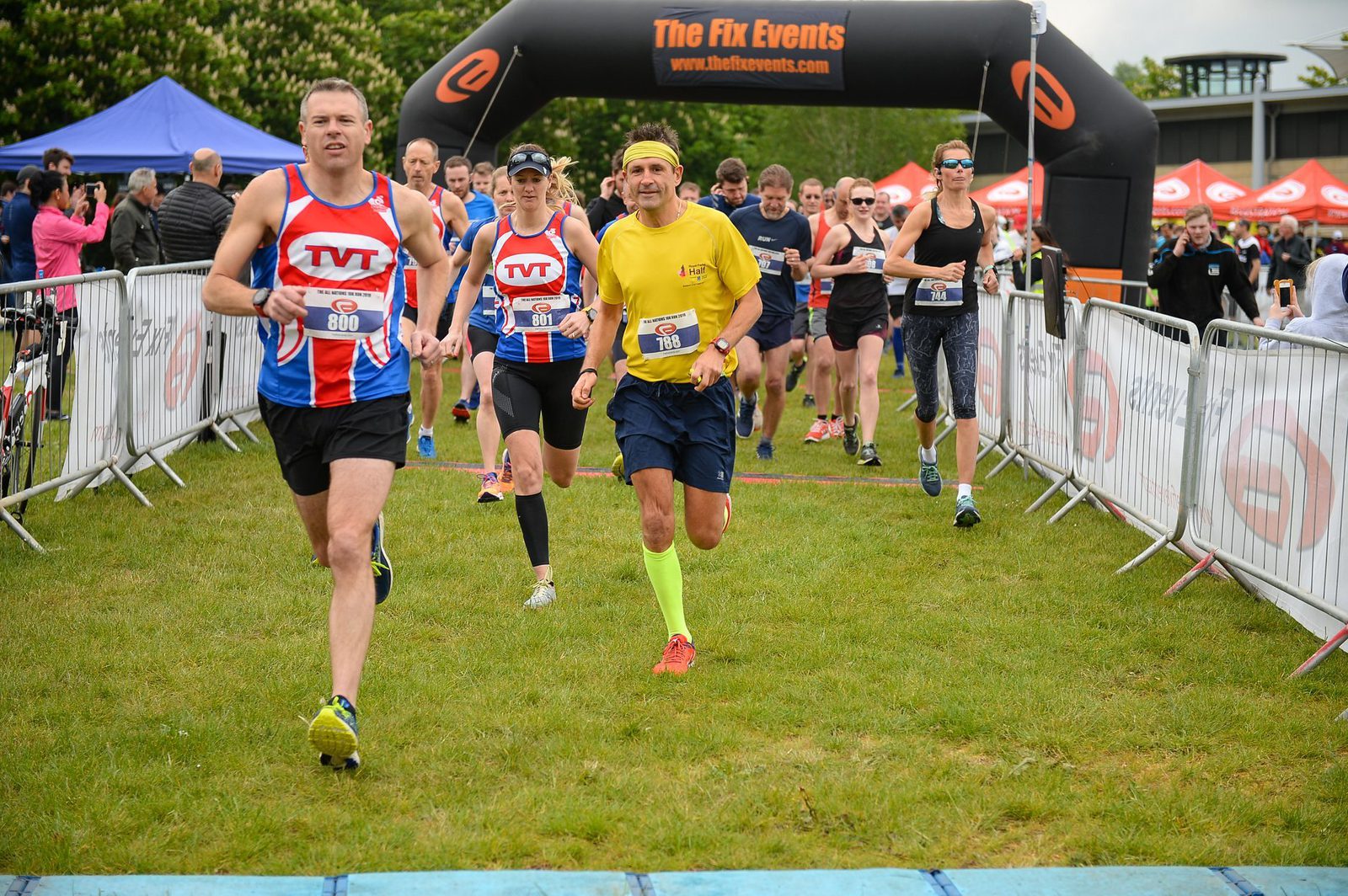 Runners participating in a race event, with some wearing bibs and branded athletic attire. They are approaching the finish line under an archway labeled "The Fix Events." The scene is outdoors with a grassy area and tents in the background.