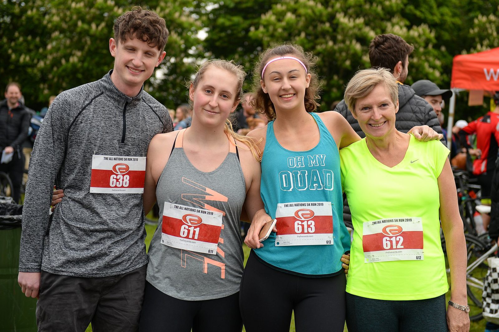 Four people are grouped together outdoors, smiling at the camera. They are wearing athletic clothing and numbered race bibs (638, 611, 613, 612). In the background, more participants and trees can be seen.