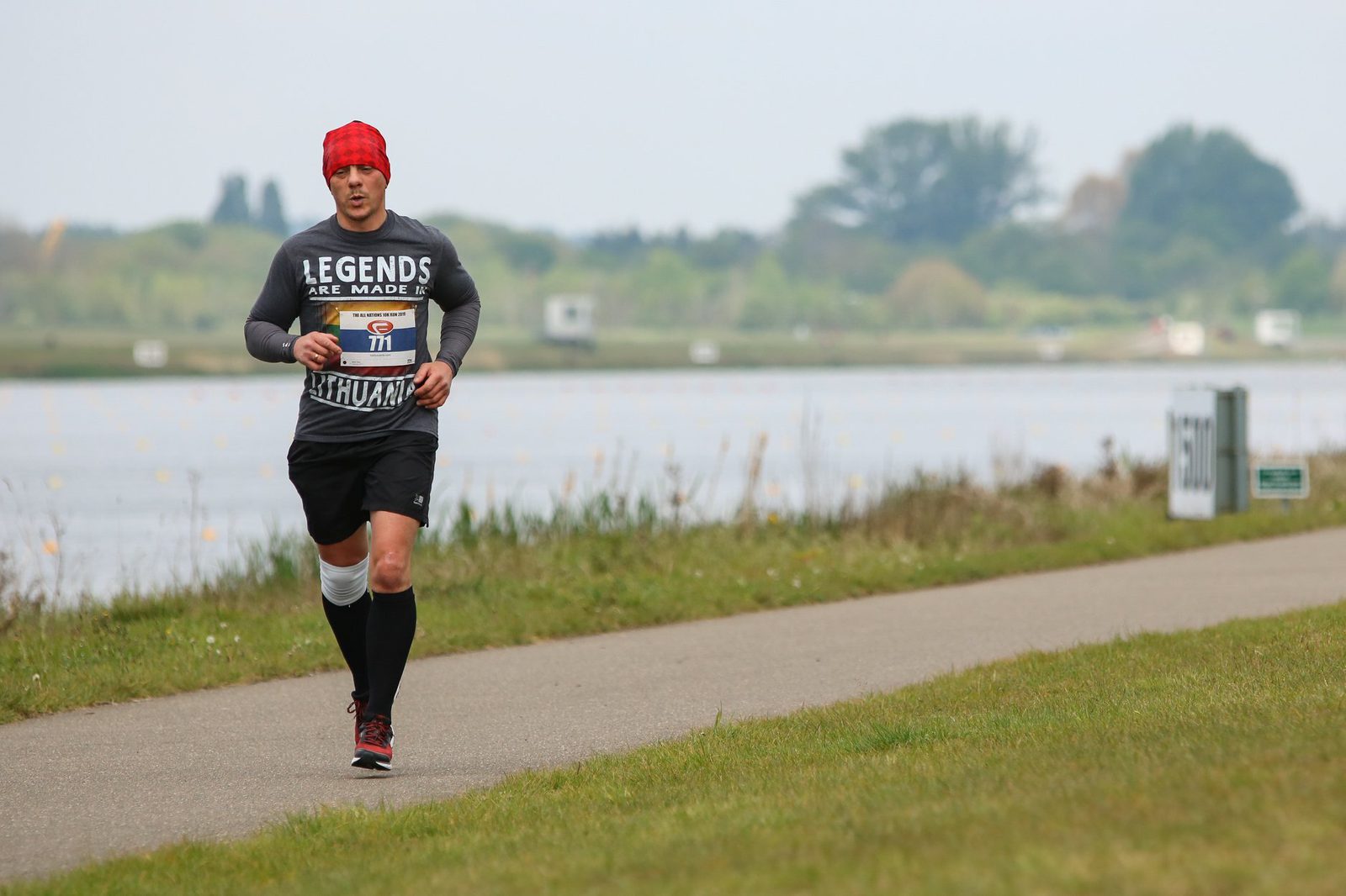A runner wearing a red headband, black shirt with "Legends are made" text, black shorts, and black knee-high socks is running on a lakeside path. In the background, green trees and a body of water are visible under a cloudy sky.