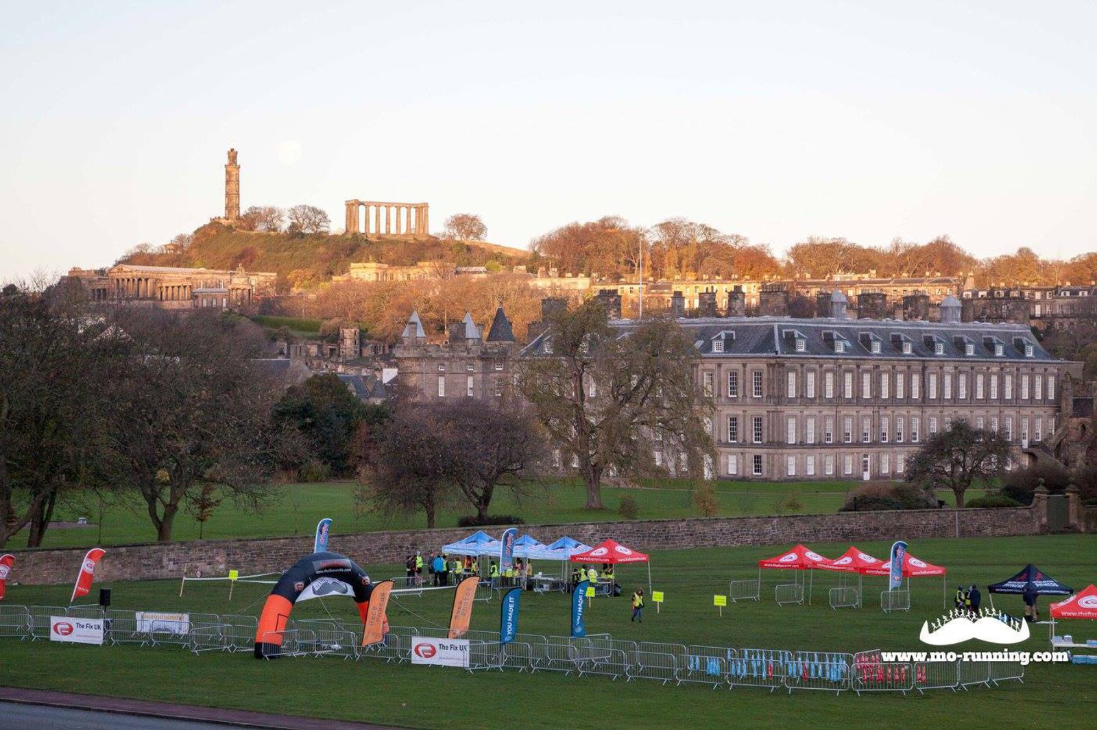 A large lawn with several blue, red, and black tents set up, flanked by metal barriers. People are scattered near the tents. In the background, a hill with the National Monument and Nelson Monument on top, set against a clear sky. A long building is visible in the foreground.