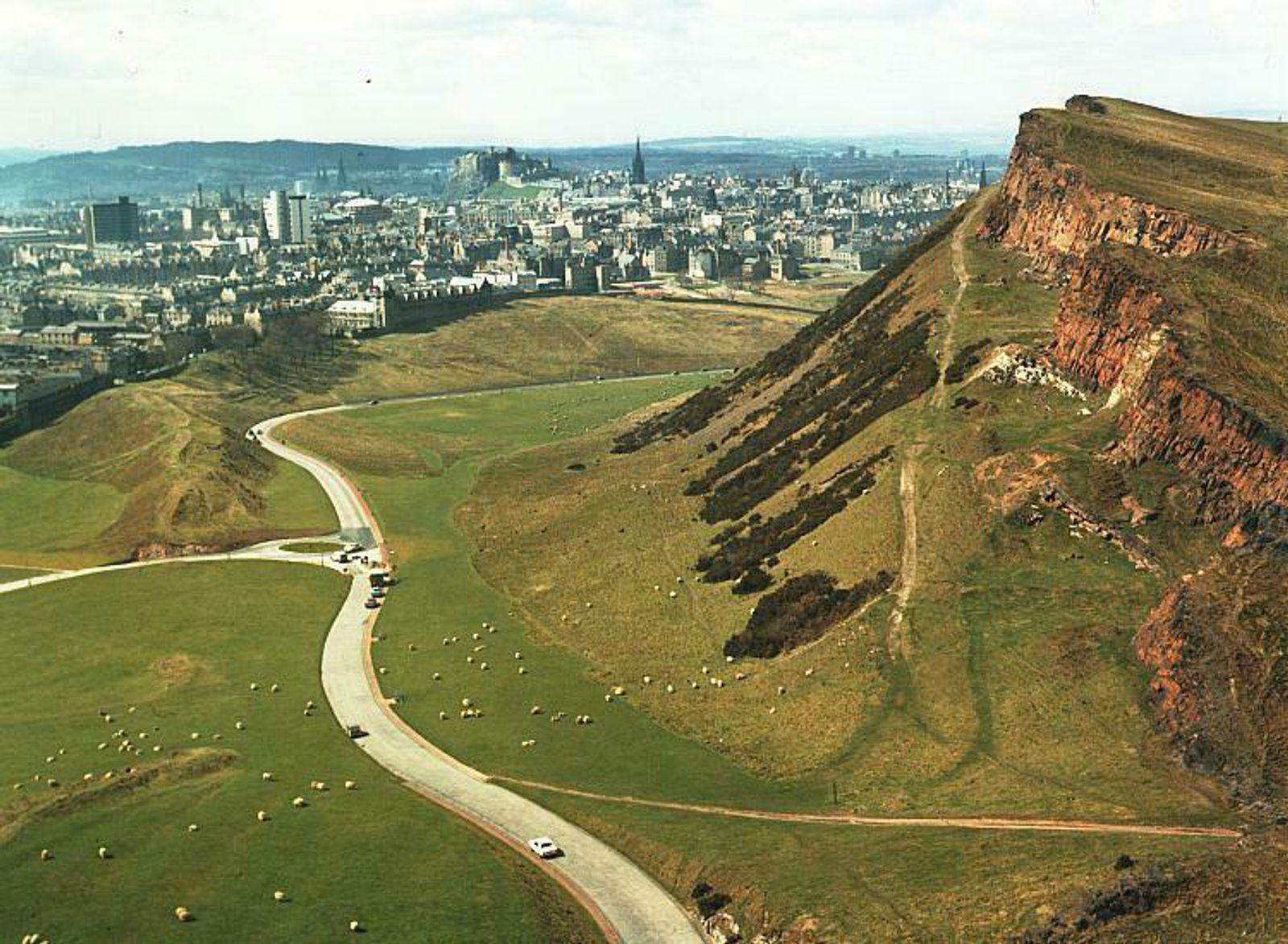 A panoramic view of a cityscape with a large, grassy hill in the foreground. The hill has a steep, rocky cliff face on the right. A winding road passes through the green landscape, and the city with numerous buildings is visible in the distance under a cloudy sky.