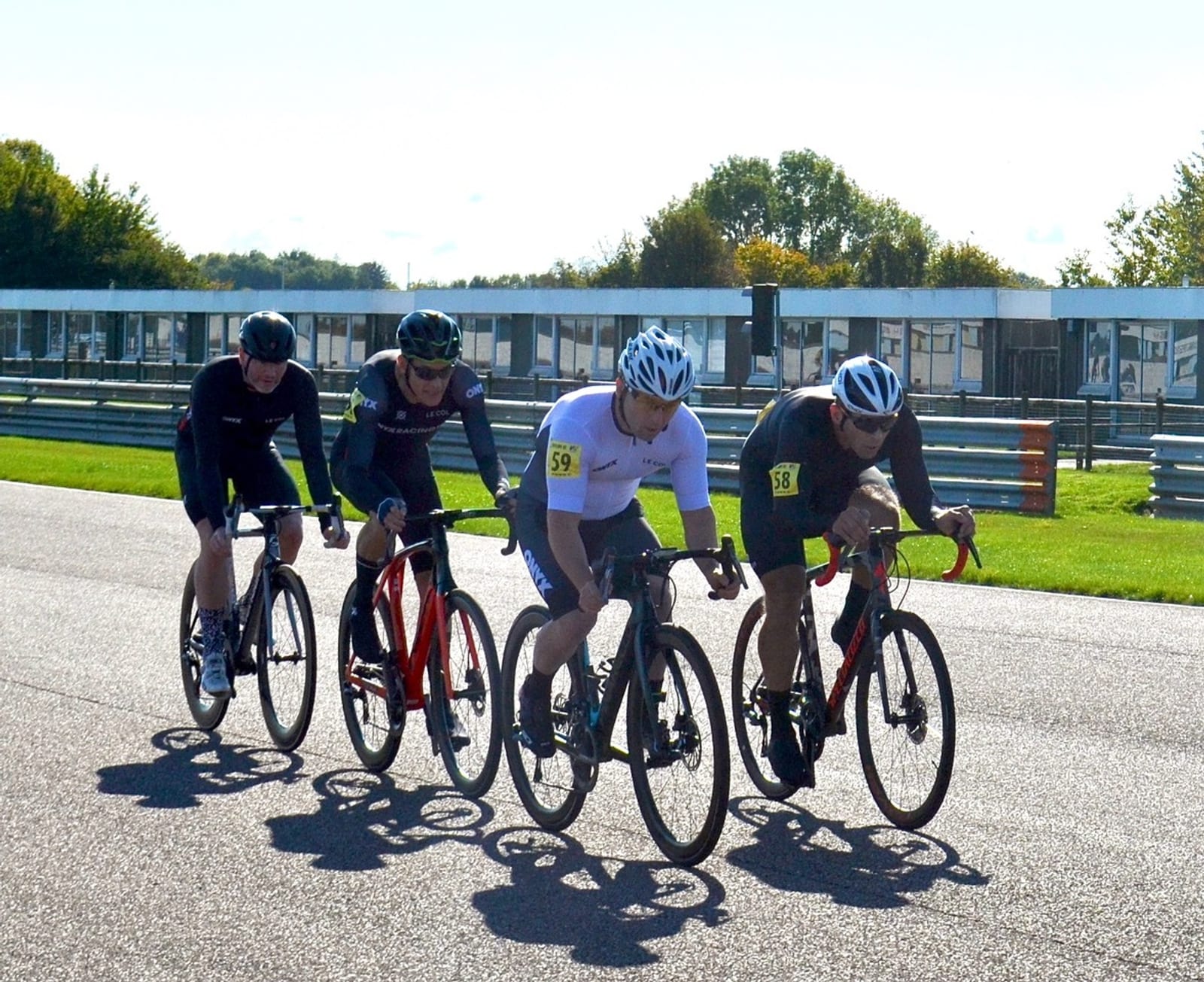 Four cyclists in racing attire and helmets ride closely together on a sunny day, possibly during a competitive event. They are on a paved track with green grass and a building in the background. Each cyclist has a number tag on their jerseys.