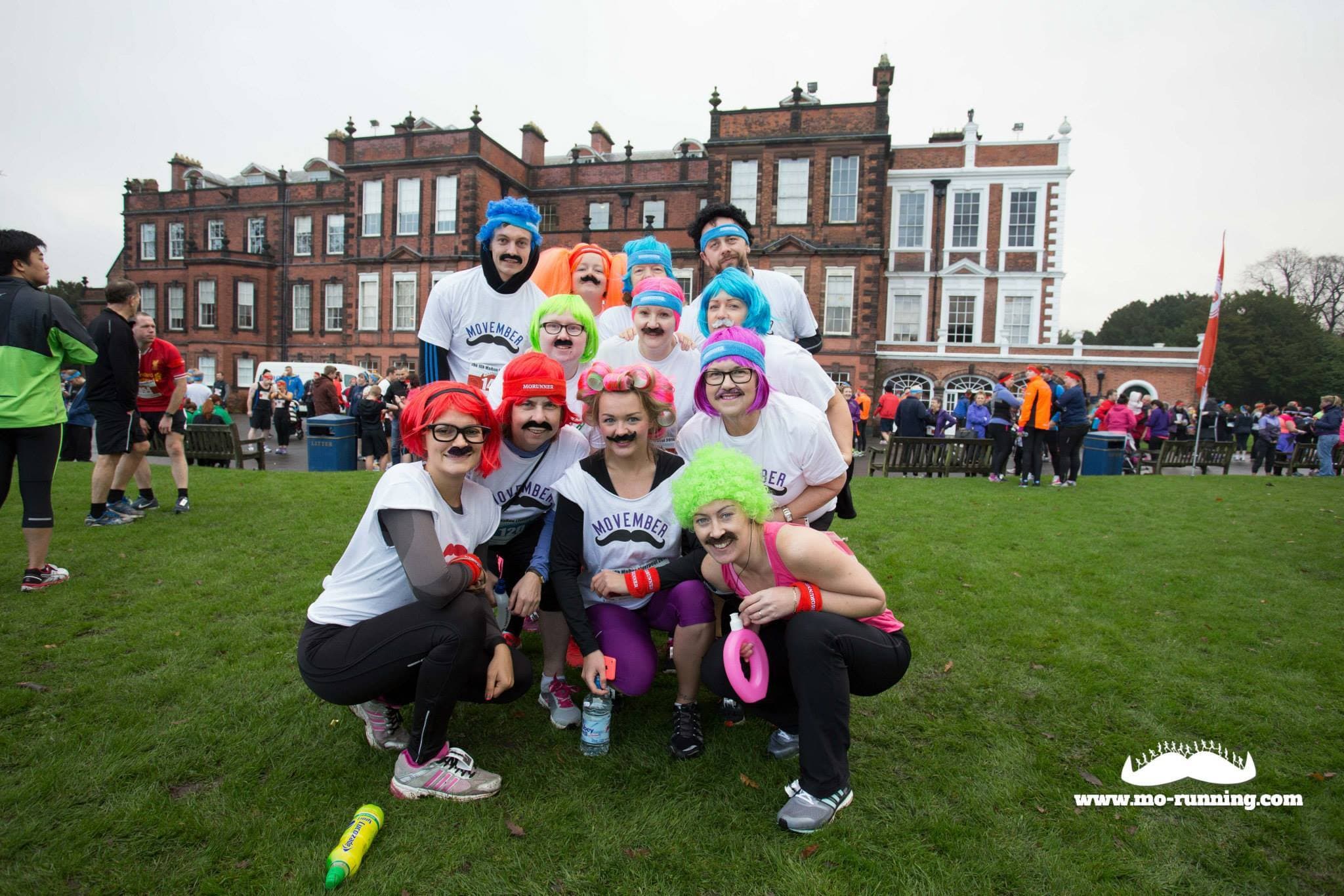 A group of people dressed in playful costumes, including colorful wigs and fake mustaches, posing for a photo in front of a large historic building. They are holding novelty items and wearing matching "Movember" t-shirts, participating in an outdoor event.