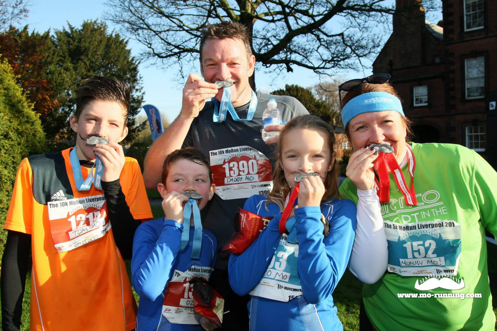 A group of five people, including adults and children, are standing outside and smiling at the camera, each biting a medal. They are wearing race numbers and assorted athletic clothing, indicating they have participated in a running event. A banner and trees are visible in the background.